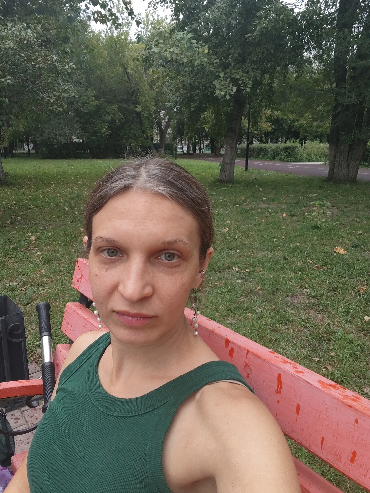 A woman with graying brown hair pulled back and light blue eyes is seated on a pink park bench in a park setting. She is wearing a green ribbed tank top and long dangle earrings with pearls. Her head is turned slightly towards the camera. In the background, trees, a path, and grass are visible. A black metal trash can is to her left.