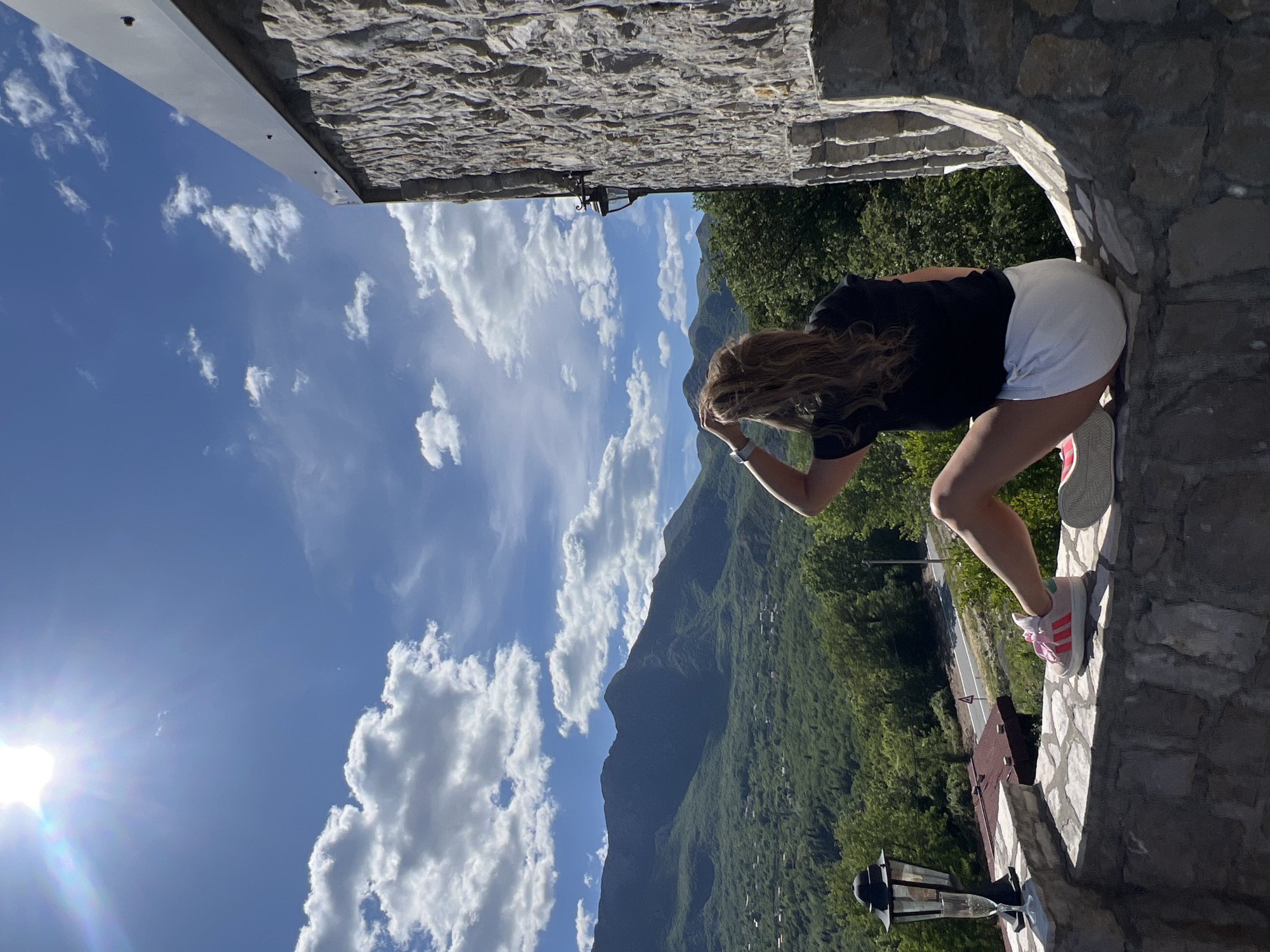The image, originally oriented horizontally (landscape) and rotated 90 degrees counter-clockwise, captures a picturesque daytime scene from an elevated stone structure in Sotonići, Montenegro. In the foreground, a person, appearing to be a woman with long, wavy light brown hair, is bent over, looking out over the expansive mountainous landscape. She is dressed in a black t-shirt, white shorts, and white sneakers with pink stripes and laces. Her left arm is raised towards her head, possibly shielding her eyes from the sun or adjusting her hair.
The immediate surroundings feature rustic stone masonry, forming a wall and an arched opening or balcony from which the view is enjoyed. A traditional black lantern with glass panels is affixed to the stone wall on the right side of the frame. Beyond this structure, a vast, verdant mountainous terrain stretches into the distance under a bright blue sky. Numerous white, fluffy clouds are scattered across the sky, with the sun prominently visible in the upper left, radiating a strong glare and indicating clear weather. A winding road can be seen snaking through the forested slopes, and some small, distant buildings or rooftops are nestled among the trees. The scene evokes a feeling of awe and appreciation for nature's beauty from a serene, elevated vantage point. There is no visible text in the image.