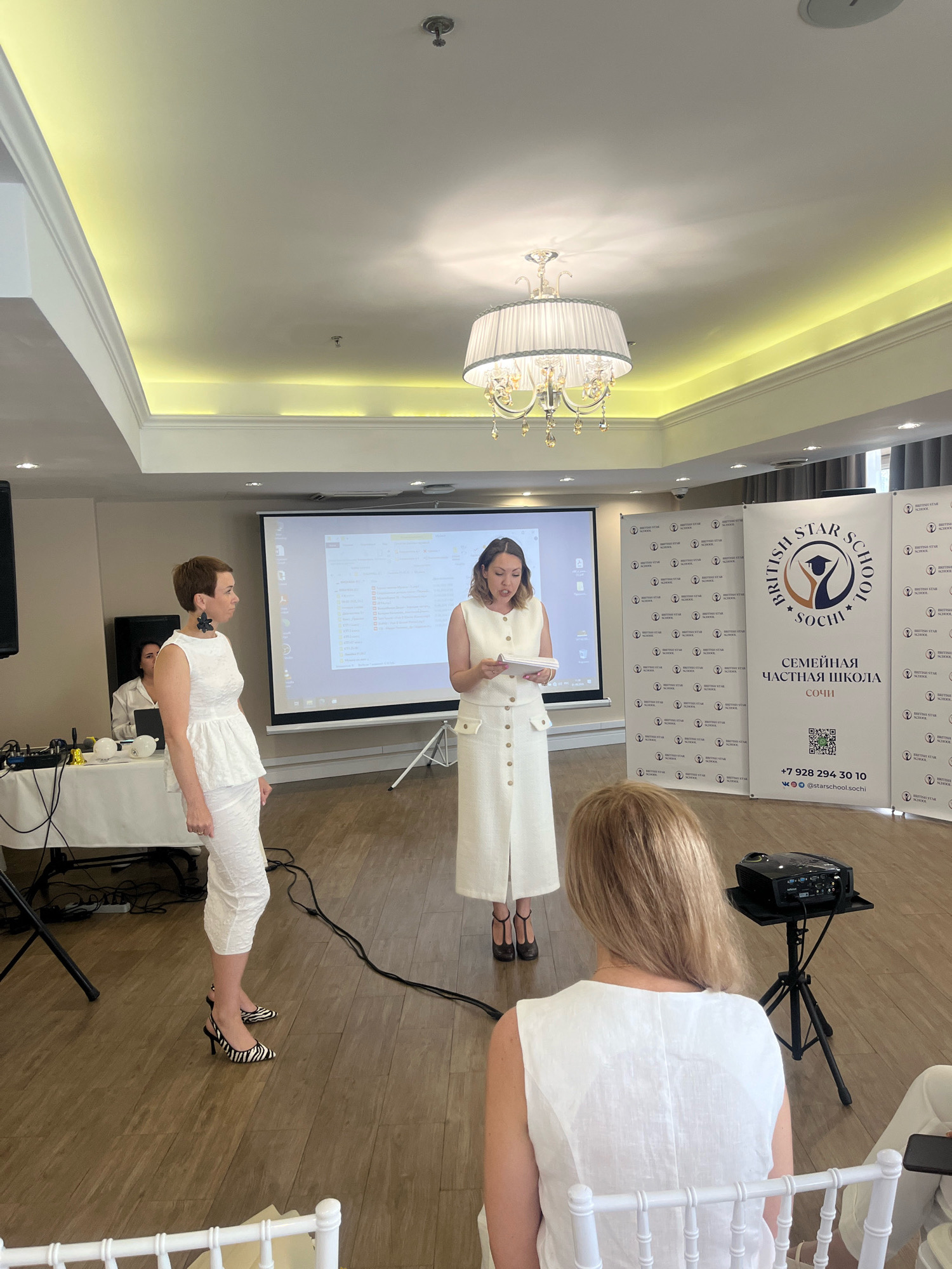 The image depicts an indoor event, likely a presentation or a gathering, at the British Star School in Sochi, Russia. Two women are the central figures. The woman on the left, dressed in a white peplum top and matching skirt, stands facing a projector screen. She has short dark hair and is wearing zebra-print high heels. To her right, a second woman, also in white attire, is holding a document and appears to be speaking. Behind the speakers, a large banner displays the "BRITISH STAR SCHOOL SOCHI" logo, along with contact information and a QR code. A projector is set up on a tripod in the foreground. The environment includes a wooden floor, a white table with equipment on the left, and a decorative chandelier hanging from the ceiling. The lighting suggests a daytime event.