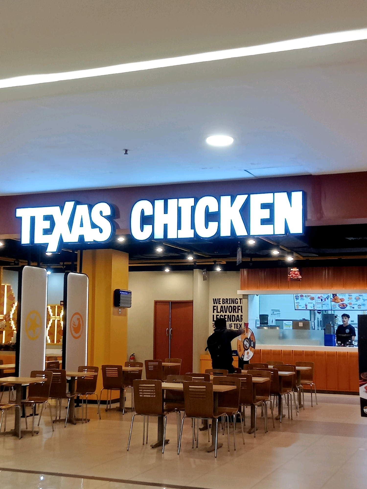 The image shows the interior of a Texas Chicken fast-food restaurant, likely located in a mall in Cirebon, Indonesia, given the context of similar locations. A large, illuminated "TEXAS CHICKEN" sign is prominently displayed on a dark-colored facade. Below the sign, there are tables and chairs arranged for dining. Two tall, white columns with orange circular logos are visible to the left. To the right, a counter area is set up with a menu board displaying food items and two employees behind it. One employee is taking an order or serving a customer, while another is visible further down the counter. A man in a dark jacket is standing near a prize wheel, appearing to be interacting with it or preparing to spin it. The restaurant has a clean, modern interior with tiled flooring. The lighting appears to be a combination of ambient ceiling lights and the bright illumination from the signage.