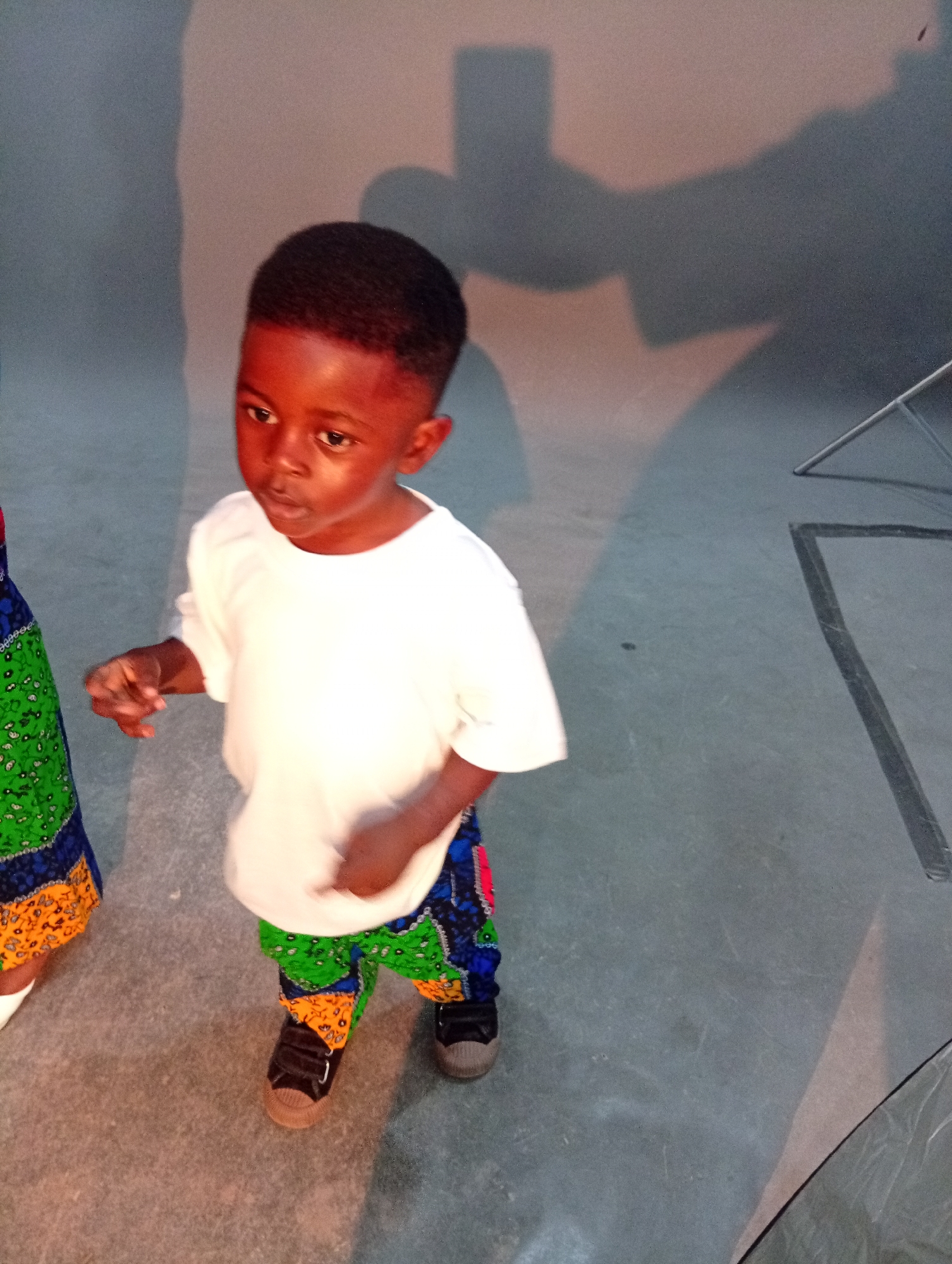 An overhead shot shows a young Black boy standing in what appears to be a studio with gray walls and a concrete floor. The boy is facing the left side of the frame with his head slightly tilted and looking down and to his left. He has short, dark hair and dark skin. He is wearing a plain white t-shirt and colorful pants with a pattern of green, blue, orange, and yellow shapes. His right hand is in the foreground, slightly blurred, as if reaching out or gesturing. He is wearing black sneakers. To the left of the boy, a person in a green and blue patterned garment is partially visible. Shadows of figures are cast on the wall behind the boy. In the background on the right, a portion of a metal stand and a dark rectangular object are visible.