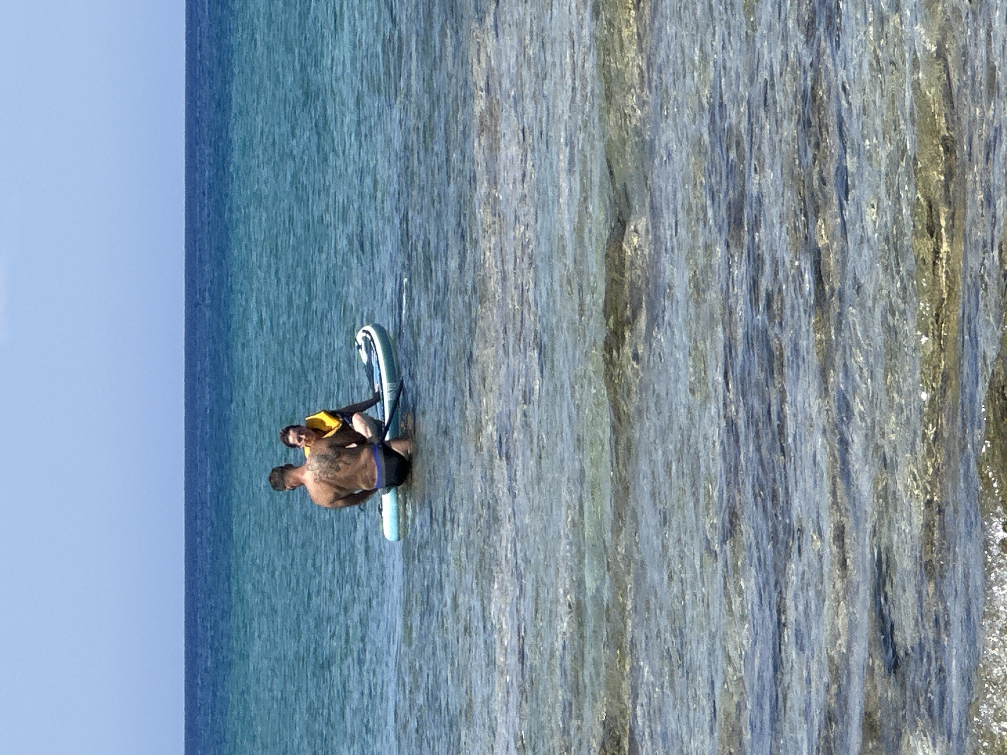 A medium shot captures two individuals seated on a paddleboard in a body of water under a clear blue sky. The water is a vibrant turquoise, transitioning to a deep blue horizon line where it meets the sky. The foreground is filled with shallow, clear water, revealing submerged rocks and sandy patches.
The person on the left, viewed from the back, is a muscular individual with extensive tattoos on their back and shoulders, wearing dark swim trunks. They appear to be looking towards the person on the right. The person on the right is facing the camera, wearing a yellow life vest over dark swimwear. They have short, dark hair and are looking at the person on the left. Both are seated on a white and blue paddleboard, with the front of the board angled towards the right. The water around the paddleboard is calm, with gentle ripples.