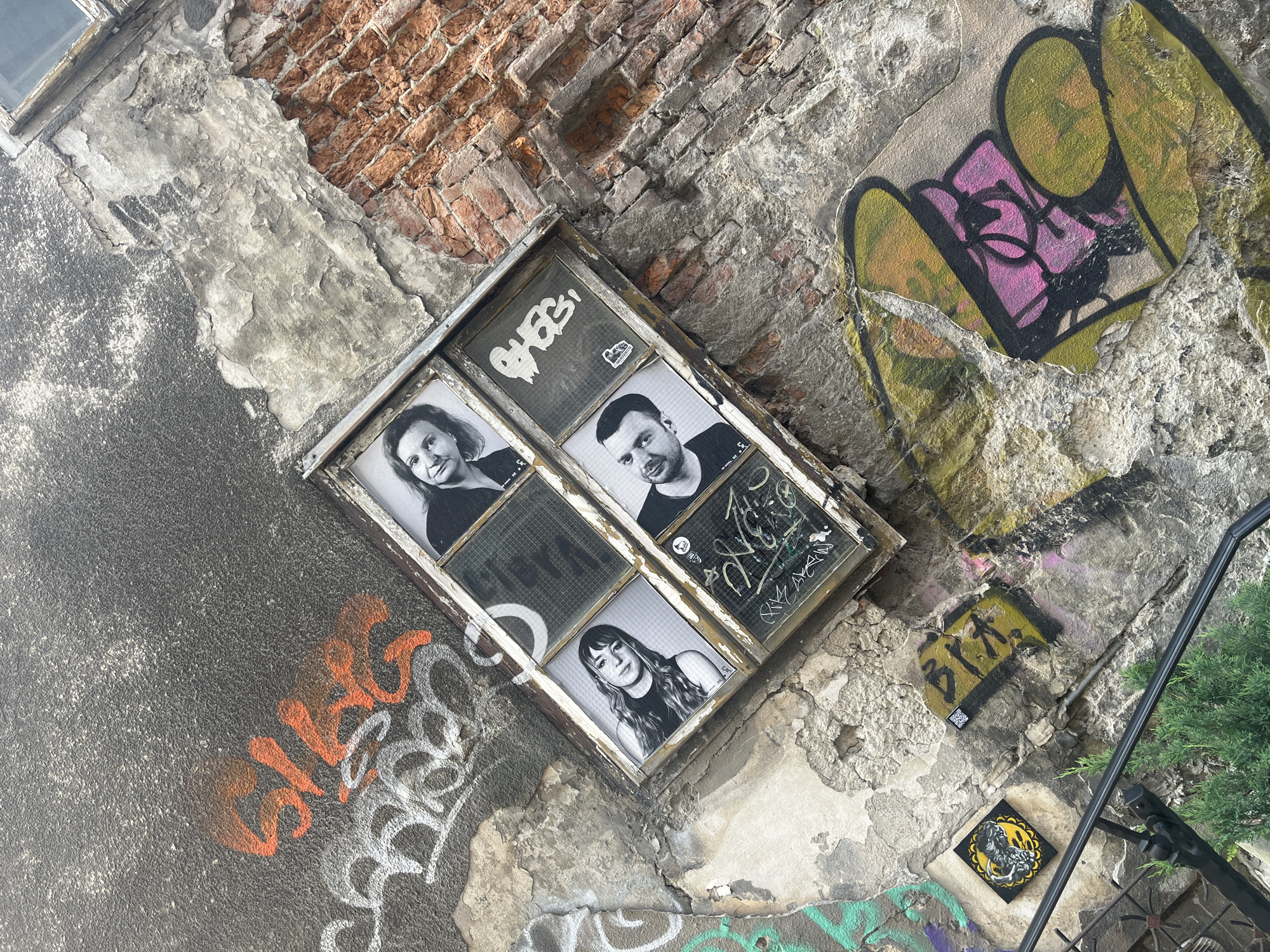 The image shows a section of an old, weathered wall in an urban environment, likely a city. The wall is a mix of rough stucco and exposed brick, with significant signs of age and neglect, including cracks, peeling paint, and moss. The wall is covered in various forms of graffiti.
Dominating the center of the image is a framed, multi-paned window or opening that has been repurposed to display four black and white photographs. These photographs, arranged in a 2x2 grid within the window panes, feature portraits of individuals. The top left pane shows a woman with shoulder-length hair, smiling slightly. The top right pane shows another woman with longer hair, also with a subtle smile. The bottom left pane features a man with short hair, looking towards the viewer. The bottom right pane shows another man with dark hair and a beard, looking slightly off-camera. Some of the window panes also have graffiti applied directly to them or to stickers placed on them.
Surrounding the window are other graffiti tags and art. To the left of the window, there is a large orange graffiti tag with white outlines. Below this, there is more white graffiti. To the right of the window, the exposed brick wall is visible, also showing signs of wear.
Further down the wall and to the left, there are smaller pieces of street art, including a square yellow and black graphic with a stylized animal or monster and a yellow rectangular tag with black lettering and a QR code. In the bottom right, a large abstract graffiti piece in shades of pink, green, and yellow is visible. A black metal railing and some greenery are present in the foreground at the bottom of the image, suggesting the wall might be adjacent to a walkway or public space. The overall impression is one of urban decay and artistic expression through graffiti and the recontextualization of found elements.