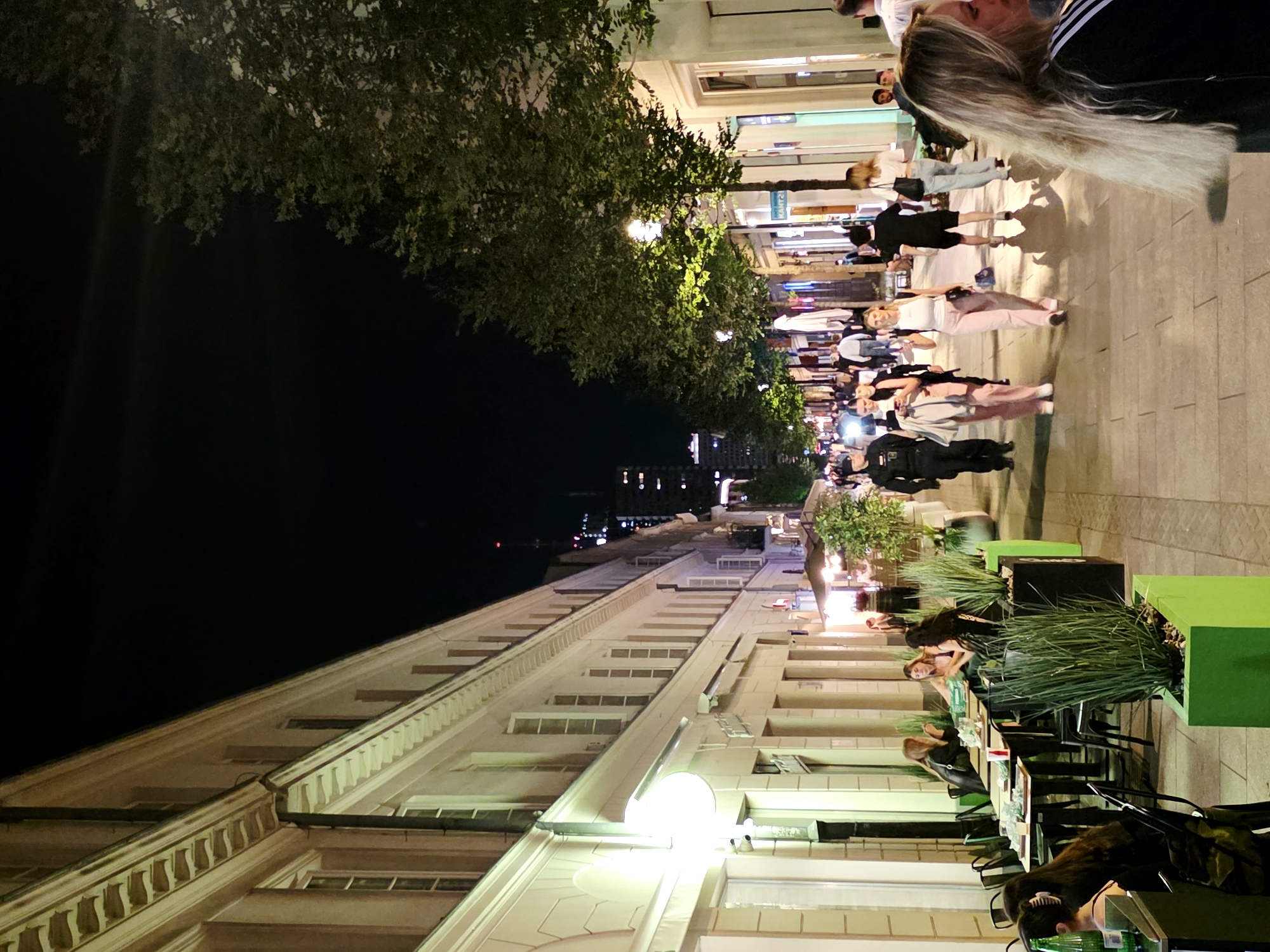 The image depicts a street scene at night in Warsaw, Poland, likely near the Palace of Culture and Science, indicated by the tall building in the background. The street is lined with buildings featuring ornate architectural details and large windows. Several people are walking along the sidewalk, which is illuminated by streetlights and lights from the buildings and outdoor seating areas of restaurants. Some individuals are seated at outdoor tables, suggesting a lively evening atmosphere. Trees are planted along the sidewalk, their leaves illuminated by the ambient light. The overall impression is of a bustling urban environment at night, with a mix of pedestrian traffic and commercial activity.