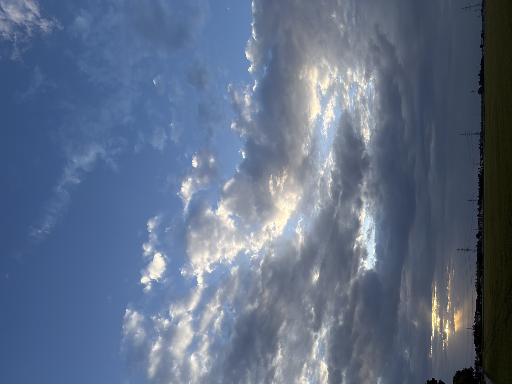 The image shows a sky full of clouds over a rural landscape. The sky is a vibrant blue with scattered white and gray clouds. Some of the clouds are illuminated by the sun, creating a beautiful contrast. Below the sky, there is a flat field of green grass. In the distance, there are a few houses and some power lines, suggesting a sparsely populated area. The sun is setting, and its rays are visible through a gap in the clouds, casting a warm glow on the landscape. The overall scene is serene and picturesque.