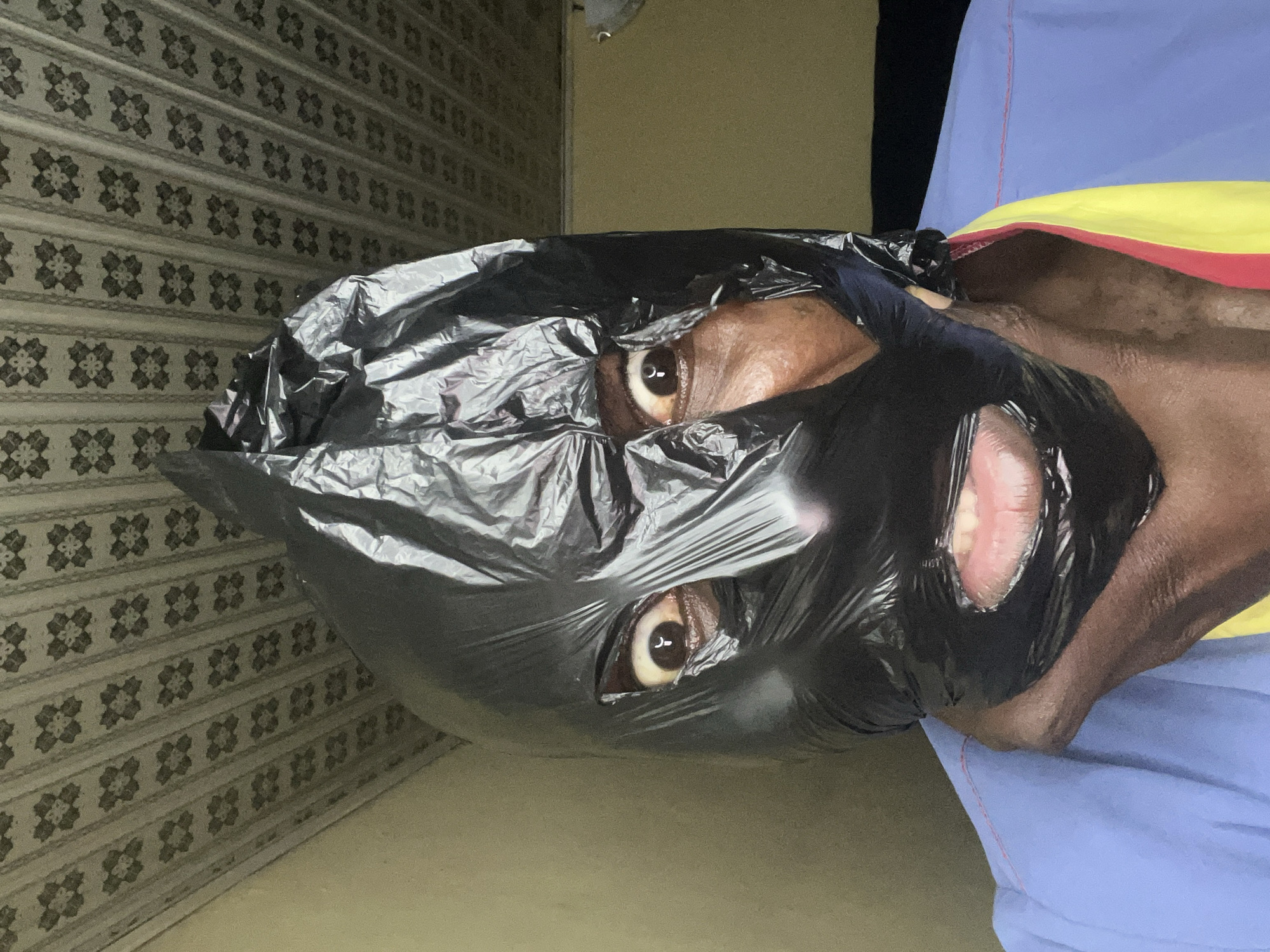 An indoor, medium close-up image captures a person, identified as being in Gura Top, Nigeria, looking directly at the viewer. The individual has dark skin and is wearing a shiny, crinkled black plastic bag or mask over their head, which covers most of their face. Cut-outs in the plastic reveal the person's wide-open eyes, both looking forward. The whites of their eyes have a yellowish tint. An opening for the mouth also shows slightly parted lips, revealing some teeth and the inner mouth. The plastic material is reflective, indicating its thin, flexible nature.
The person is wearing clothing that includes a blue fabric, possibly a shirt or tunic, with a distinctive yellow and red stripe visible beneath it. The background consists of a wall partially covered in patterned wallpaper or a decorative surface featuring repeating geometric or floral motifs in shades of grey and white. Above this, a plain, lighter-colored wall is visible. No specific activity is depicted beyond posing for the photograph. There are no visible location cues within the image itself to suggest Gura Top, Nigeria, and no text is present. The lighting appears to be artificial, and no specific time of day or weather conditions can be inferred.