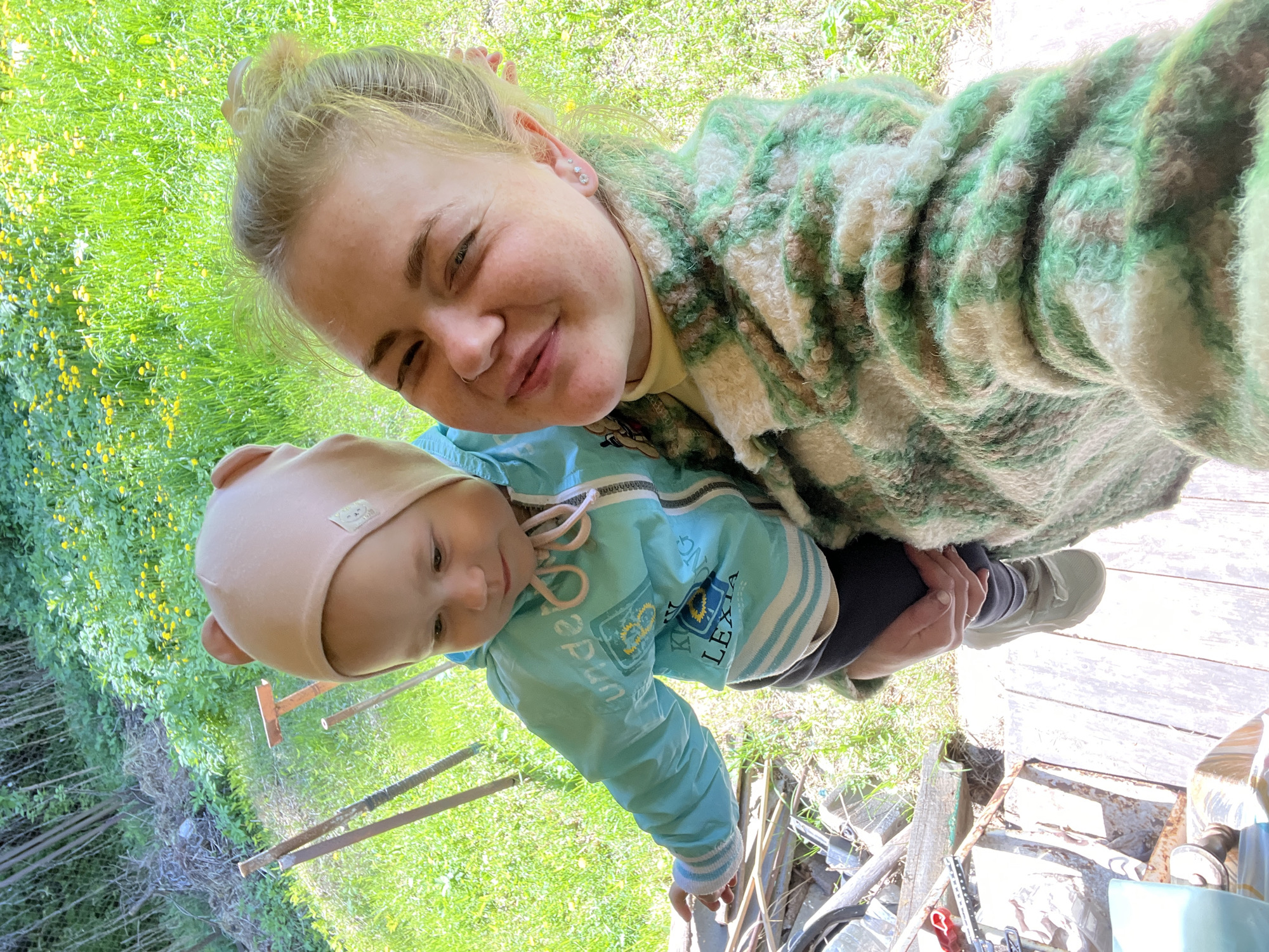 A woman is holding a baby in an outdoor setting. The woman has blonde hair pulled back in a bun and is wearing a fuzzy, green and white jacket. The baby is wearing a light blue jacket and a tan hat with bear ears. They are standing on a wooden deck, and there is grass and foliage in the background. There are tools and other objects scattered around the deck. The location is Chaikovsky, Russia.