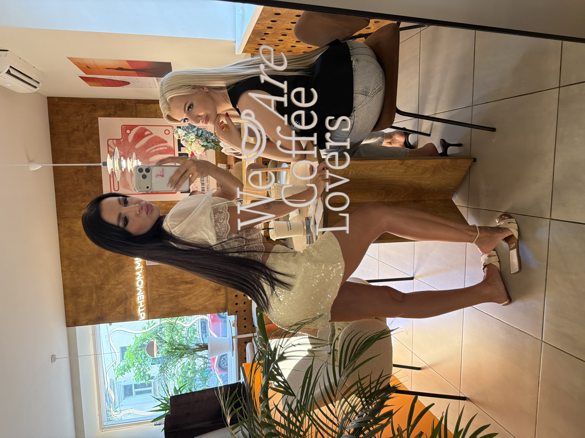 In Novaya Usman', Russia, two women are pictured inside a cafe. The woman on the left, with long dark hair, is taking a selfie with her phone, wearing a white lace top and a sequined skirt. The woman on the right, with blonde hair, is looking at the camera, wearing a black sleeveless top and jeans. They are seated at a wooden table, with coffee cups and other items on it. The cafe has a modern interior, with warm-toned walls and potted plants. The overall scene suggests a casual, social gathering between friends.