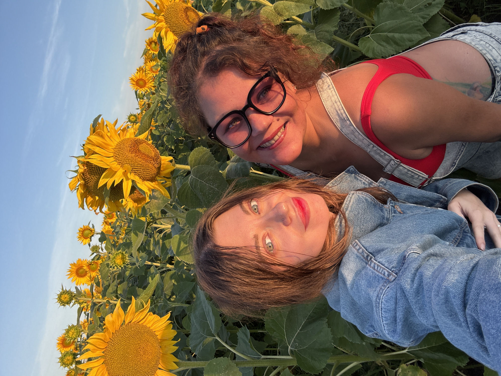 Two women are posing for a selfie in a field of sunflowers. The woman on the left has brown hair and is wearing a denim jacket. The woman on the right has curly brown hair and is wearing glasses and denim overalls over a red top. The sunflowers are tall and yellow, and the sky is blue. The image appears to have been taken during golden hour, as the lighting is warm and soft.
