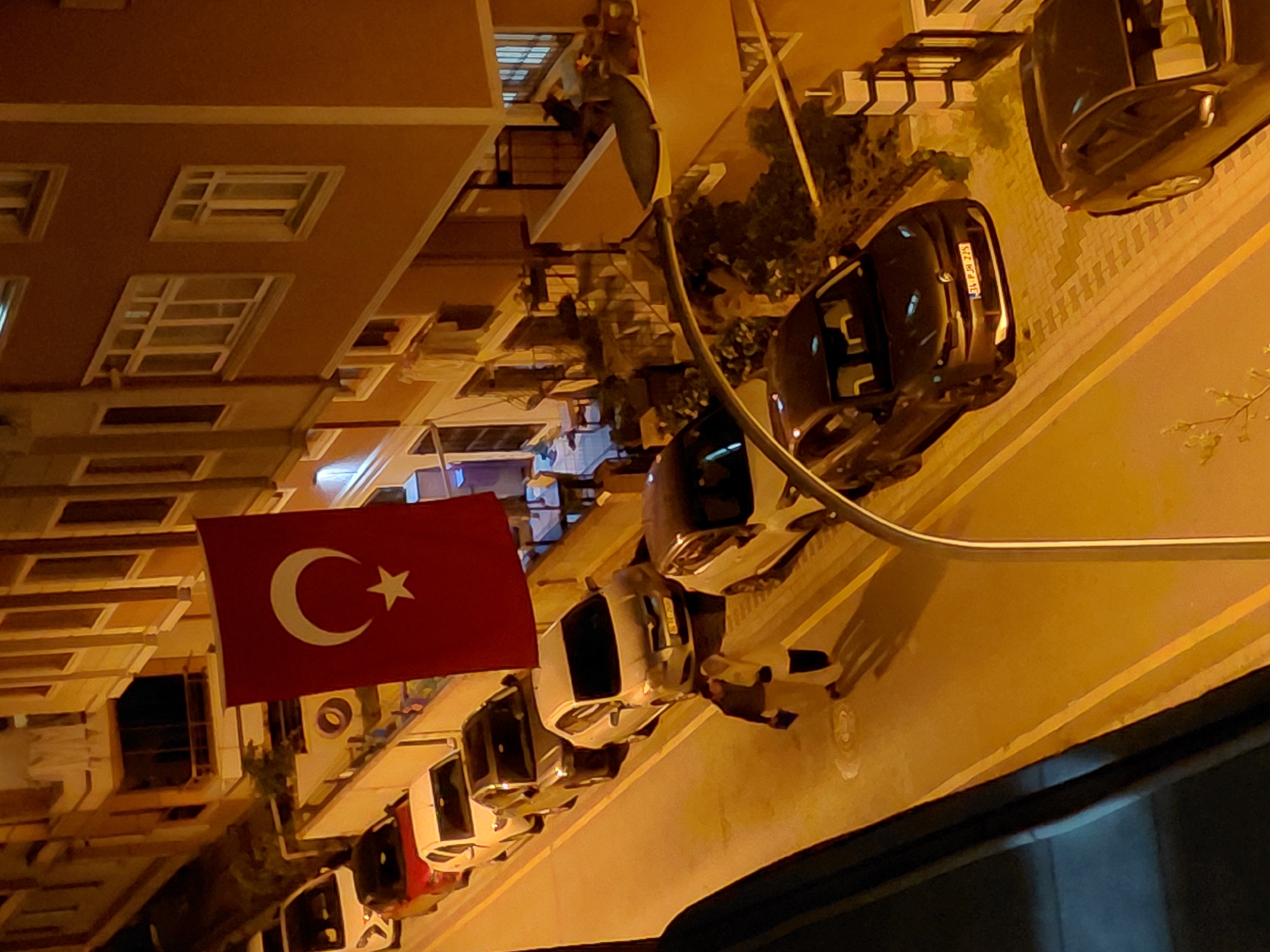 The image captures an urban street scene at night, viewed from an elevated vantage point. On the left, a multi-story residential building with a light brown facade is prominent, featuring numerous windows with white frames and balconies. Some balconies are adorned with potted plants.
A large, red Turkish flag, bearing a white crescent moon and star, hangs vertically from a balcony on the residential building, signifying the country of origin.
Along the paved street, which runs from the mid-ground towards the bottom right, several cars are parked parallel to the building. The vehicles include a mix of dark and light-colored sedans and possibly SUVs. One black car in the foreground has its license plate clearly visible, displaying "34 PJA 225". The "34" prefix on the license plate indicates the car is registered in Istanbul, which aligns with the provided location context of Üsküdar, a district within Istanbul, Turkey.
The scene is illuminated by artificial streetlights, casting a warm, yellowish glow across the buildings and the street. The time of day is clearly night due to the dark sky and reliance on artificial lighting. No people or specific activities are visible, presenting a still, quiet depiction of a residential street. The image has a slightly tilted composition.
