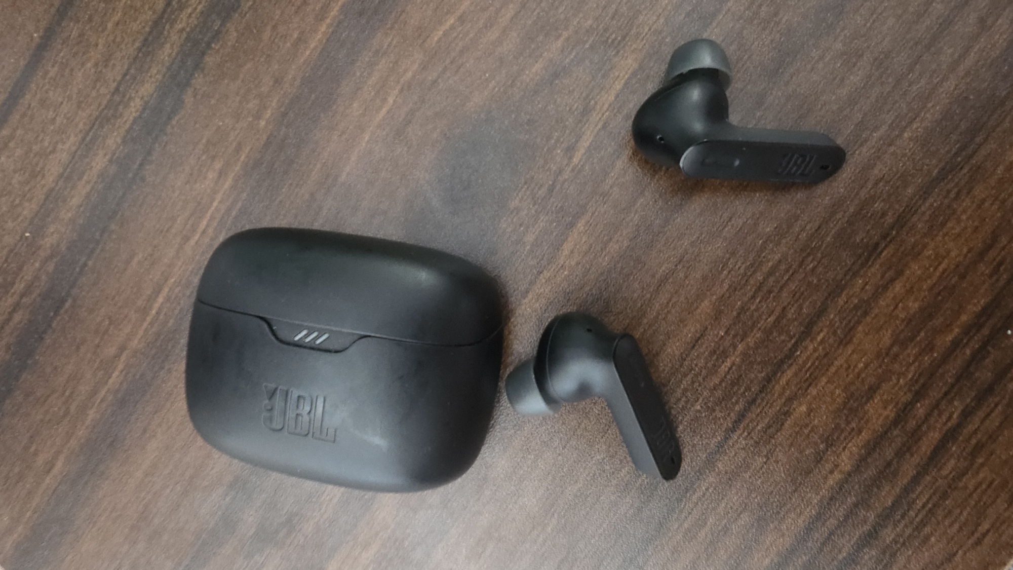 The image shows a black JBL charging case and two black JBL wireless earbuds placed on a wooden surface. The charging case is in the upper left portion of the image, with the JBL logo embossed on its surface. One earbud is positioned below and to the left of the charging case, while the other earbud is to the right and below the case. The earbuds and case appear to be clean and in good condition. The wooden surface has a visible grain pattern. The image is taken from a top-down perspective. There is no information provided about the location in the city of Bad Kreuznach, Germany, as the image itself does not contain any such contextual clues.