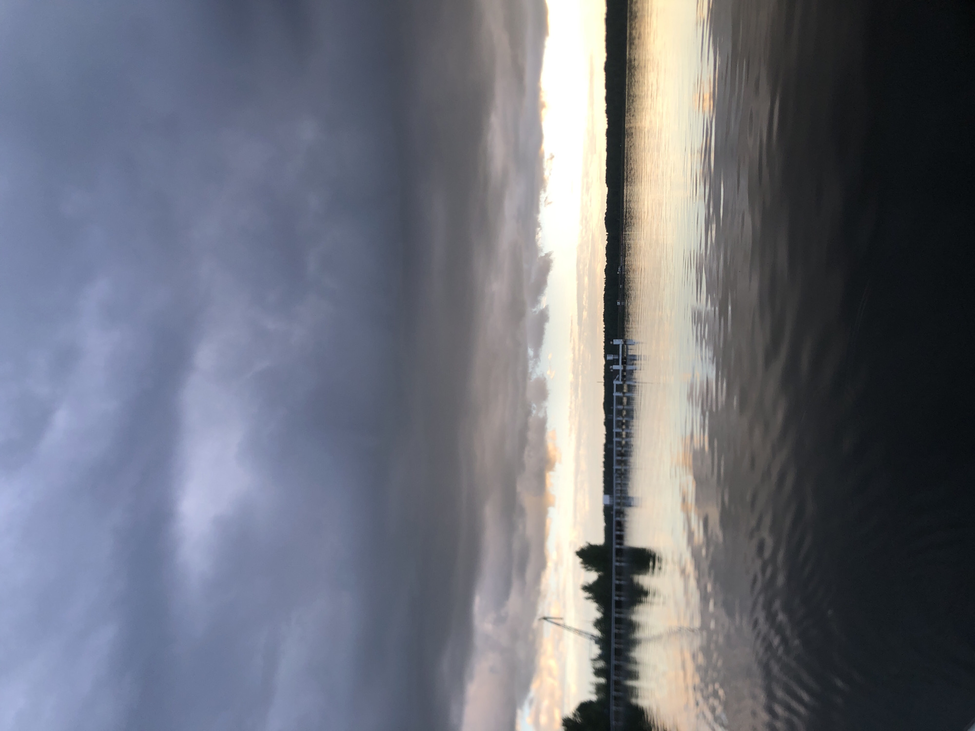This is a landscape photograph taken in Svirstroy, Russia, depicting a view of a lake at dusk. The image is oriented vertically, with the sky occupying the left side and the water the right.
The sky is dramatically overcast with heavy, dark gray clouds dominating the left half of the frame. Towards the center and right, the clouds break to reveal a bright, golden sunset. The sun itself is not directly visible but its light diffuses through the clouds, creating a luminous horizon.
The water in the foreground is calm, with gentle ripples reflecting the colors of the sky. The right side of the image shows the water in darker tones, suggesting either deeper water or less light penetration. A long, wooden pier extends into the water from the far shore, with a few small structures and what appear to be boats docked. Silhouetted trees line the distant shoreline, their shapes softened by the dim light. The overall mood of the image is serene, with a sense of approaching evening and perhaps a storm passing by. The time of day is clearly sunset, indicated by the dramatic light and the color of the sky. The location cues of a lake and pier are consistent with a lakeside setting. There is no visible text in the image.