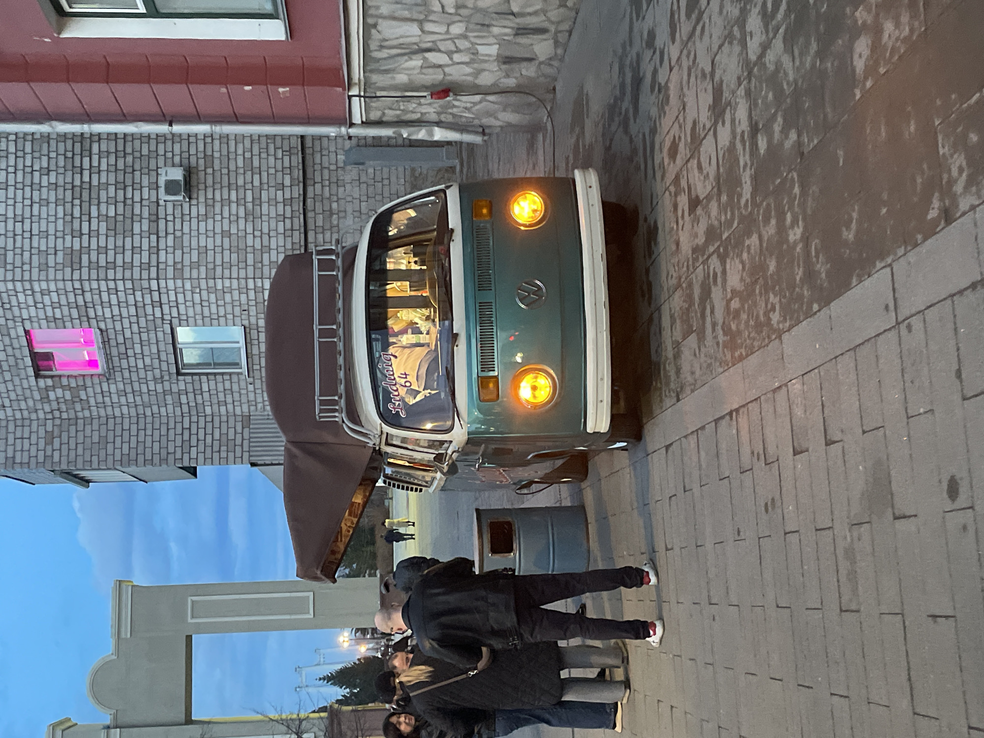 The image depicts a street scene, likely at twilight, in the city of Krasnoyarsk, Russia. A vintage Volkswagen van, painted in green and white with headlights illuminated, is parked on a stone-paved sidewalk beside a stone building with pink lighted windows. A group of people, including one in a black leather jacket, is gathered near the van, appearing to be engaging with it. An awning extends from the side of the van. A gray trash can is situated nearby. A light blue sky is visible behind a city gate, with a silhouette of trees visible in the background. The overall impression is of a street gathering around the VW van.