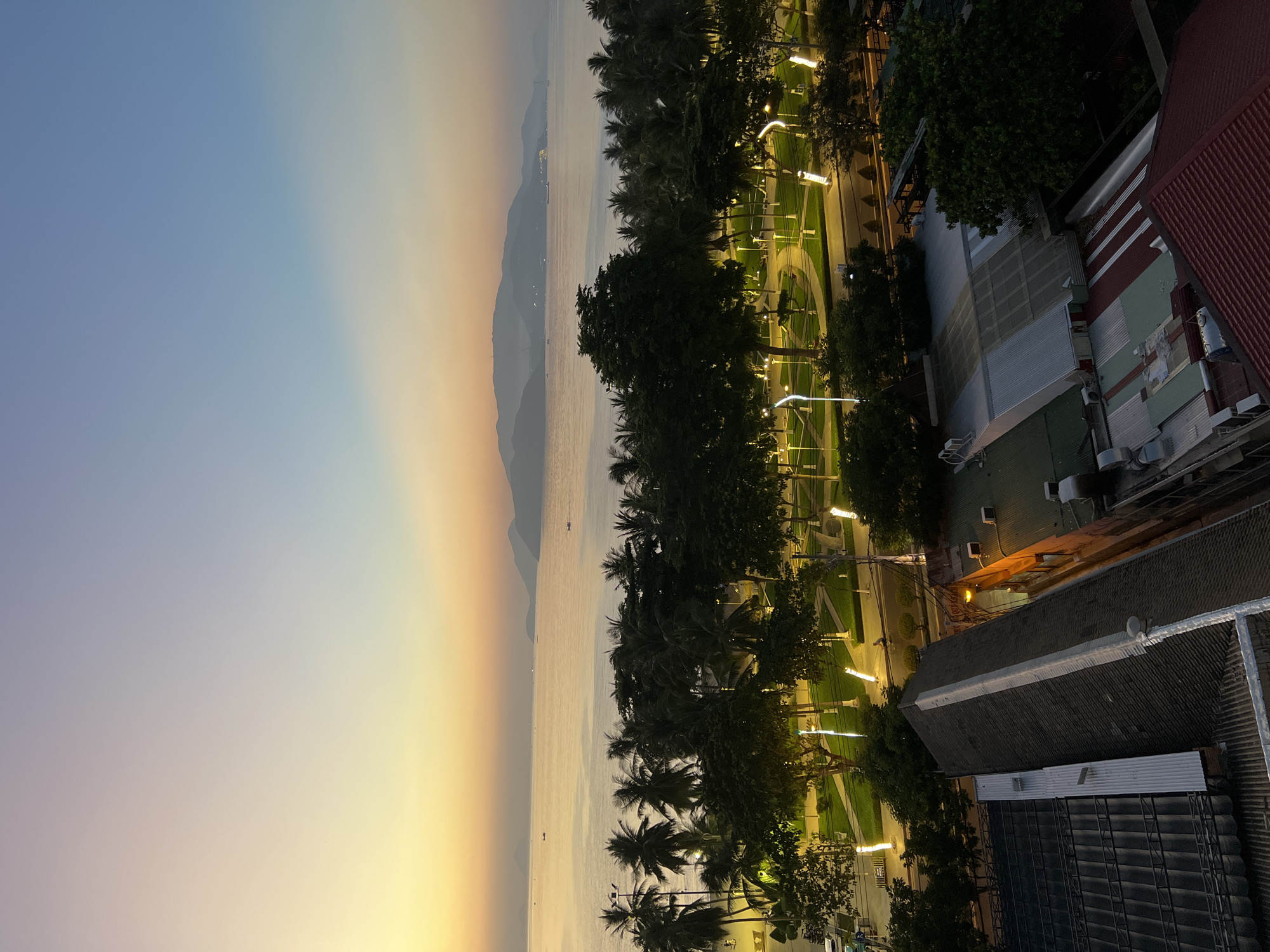 This aerial view captures a coastal city scene in Nha Trang, Vietnam, during the soft light of dusk or dawn. The horizon is dominated by a tranquil ocean that transitions into a hazy sky painted with shades of yellow, orange, and pale blue. In the distance, a chain of mountainous islands is silhouetted against the sky.
A strip of palm trees lines the shore, separating the ocean from a well-lit park area. This park features manicured green lawns, walking paths, and numerous modern, white lampposts that cast a warm glow, illuminating the space. Several figures can be seen walking or cycling within the park. A street runs alongside the park, with a few vehicles visible.
Beyond the park and street, the roofs of several buildings are visible. These range from residential structures with dark, angled roofs to commercial buildings with flatter, corrugated metal coverings. Air conditioning units are mounted on some of the buildings, and lights from windows suggest activity within. The overall impression is of a peaceful urban coastline bathed in the gentle light of twilight.