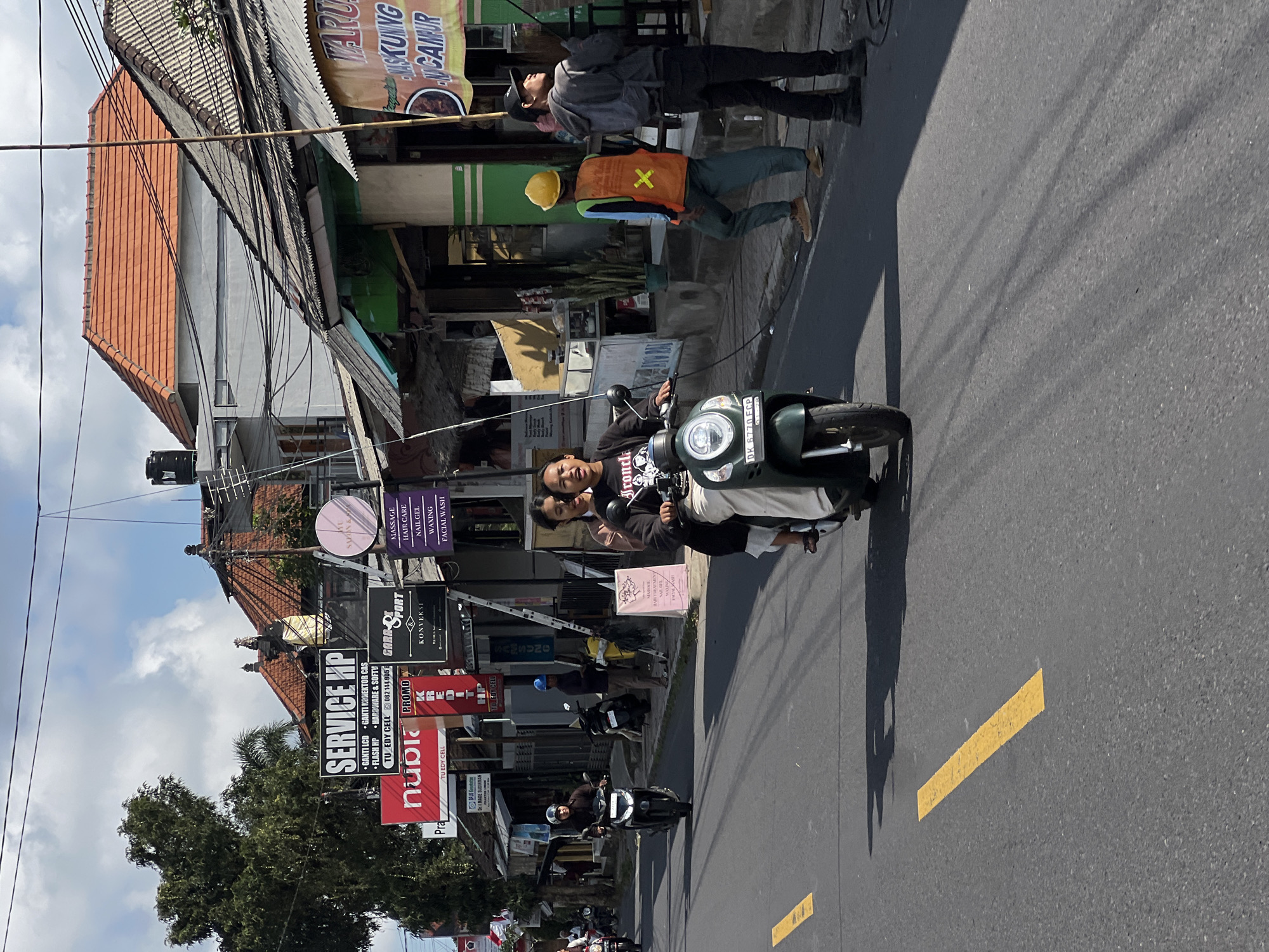 The image depicts a street scene in Tampaksiring, Indonesia, under a partially cloudy sky. In the foreground, a green scooter with two occupants, a woman driving and a passenger, travels on the right side of the road. The driver wears a black t-shirt with white lettering and the passenger wears a light-colored top and white pants. The scooter has a license plate reading "DK 6770 FCP." Yellow road markings are visible on the asphalt.
Along the left side of the street, a row of businesses with signs is visible. One prominent sign reads "SERVICE HP" with smaller text indicating phone repair services. Other signs are for "nUbia" and "PROMO."
On the right side of the street, a building with an orange tiled roof is prominent. In front of this building, several individuals are present. One person wears a bright orange vest with reflective stripes and a yellow hard hat, suggesting construction or maintenance work. Another person is wearing a grey hoodie and black pants. They appear to be observing or working on something off-camera. A banner with Indonesian text, possibly advertising food or services, hangs from one of the storefronts. The architecture features traditional Balinese elements like ornate roof structures. The environment suggests a bustling commercial area with overhead electrical wires crisscrossing the sky.