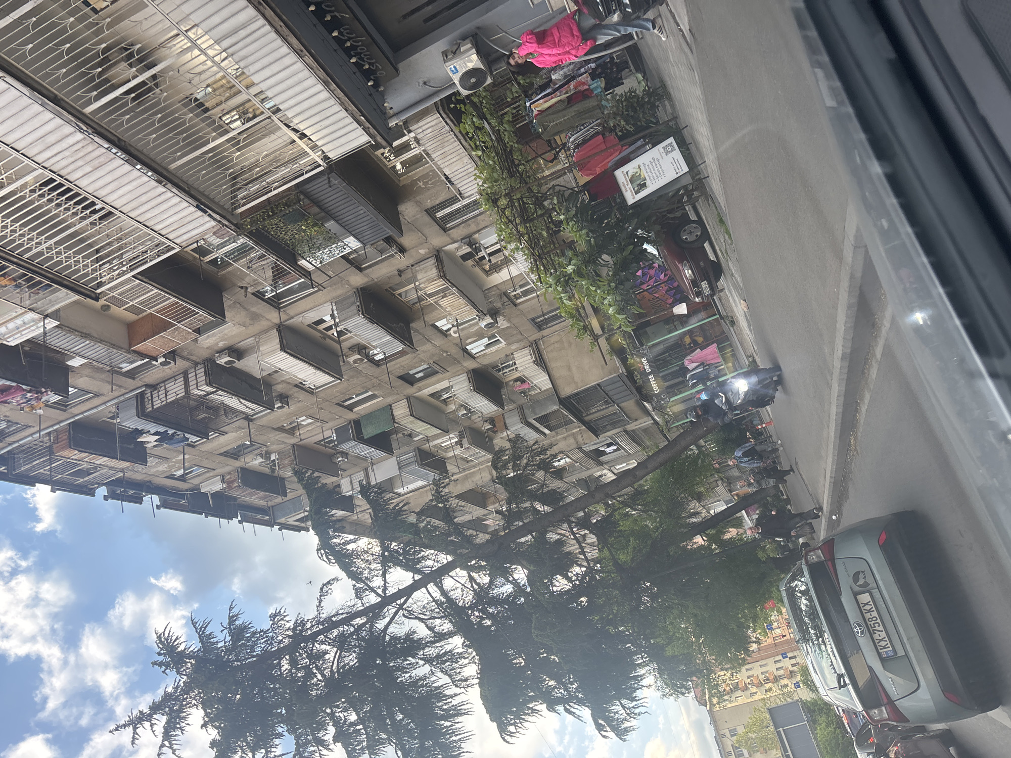 The image presents a daytime urban street scene in Tbilisi, Georgia, captured from a slightly elevated vantage point looking across a street and towards a multi-story residential and commercial building. The sky overhead is partially cloudy.
On the left, a tall, concrete-faced building dominates the frame, featuring numerous balconies and windows. Many balconies are enclosed with various materials, while others are open, displaying colorful laundry hanging, indicative of residential living. Air conditioning units are visible on the building's exterior. At street level, there are storefronts and possibly commercial establishments.
A paved street runs horizontally across the mid-ground, with a sidewalk bordering it on the right, lined with mature trees with dense green foliage. Several vehicles are visible; most notably, a light blue Toyota sedan with the Georgian license plate "KX-236-KX" is parked near the foreground on the right. Another, darker car is partially visible further down the street, and a reddish car is parked on the left side. Motorcycles or scooters are also parked along the sidewalk.
A few people can be seen in the scene. One individual wearing a bright pink jacket stands near a shop entrance on the left, while others are more obscured, engaged in daily activities like walking or sitting near businesses.
Visible text includes Georgian script on the upper part of the building and on a white informational sign positioned on the sidewalk. A partially visible sign in the bottom right corner of the image suggests "COFFEE DRIVE." The overall impression is that of a bustling, lived-in urban environment.