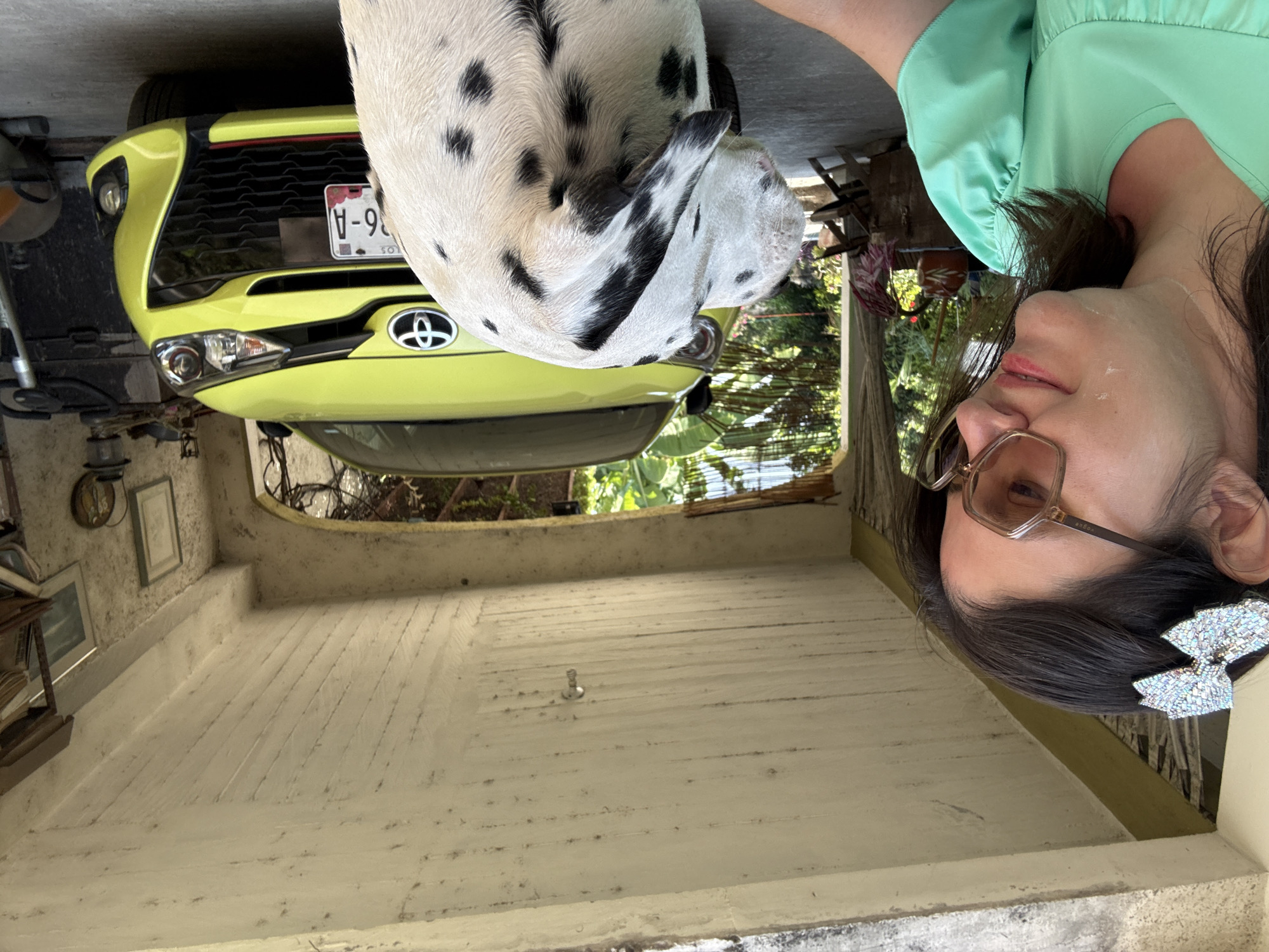 A slightly tilted, medium shot shows a woman with brown hair, glasses, and a sparkly hair bow, a Dalmatian dog, and a light yellow car inside a garage-like structure. The woman is wearing a light green shirt and faces the camera at the upper right of the frame. A Dalmatian with curled up is partially visible next to her, on the upper center of the frame, with its head facing to the right. The car, parked inside the garage, is at the upper left and angled to the left. The car has a visible license plate with the marking "V-96-A". The garage-like structure has white walls. The background shows greenery and hints of the outside environment. This scene is likely in Xochitepec, Mexico.