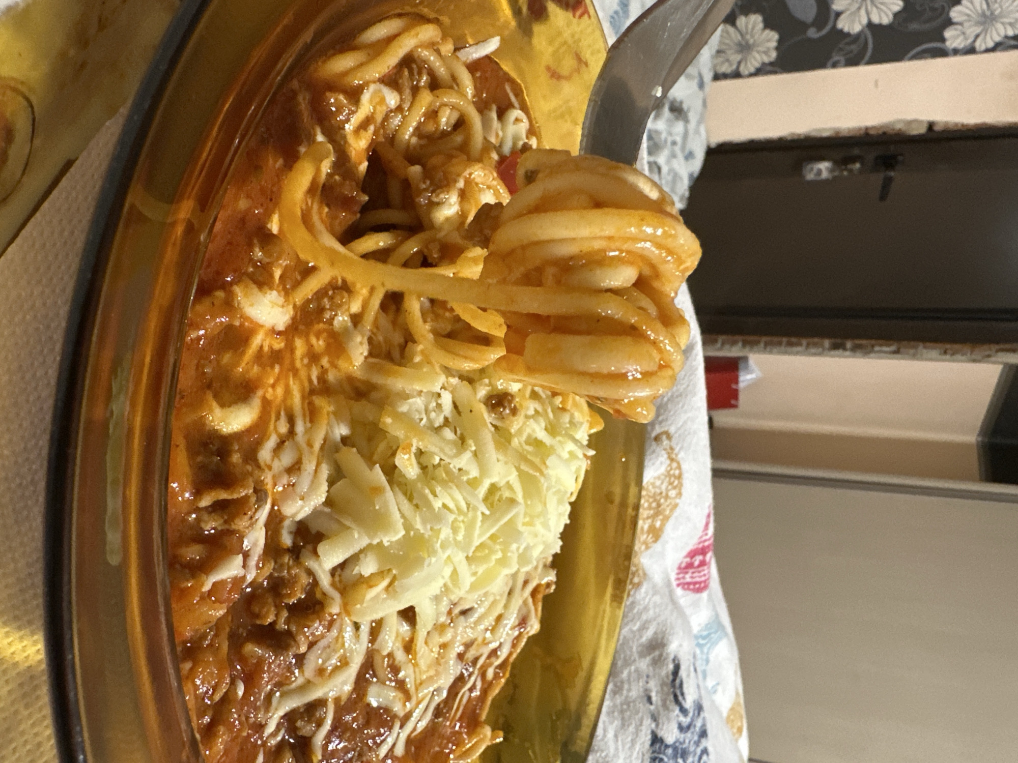 The image depicts a plate of spaghetti with meat sauce and shredded cheese. The spaghetti is being lifted with a fork, revealing the sauce and cheese underneath. The plate is gold colored and sitting on a patterned cloth. The background includes a kitchen counter and cabinets. There is no evidence to suggest the location is Sofia, Bulgaria.