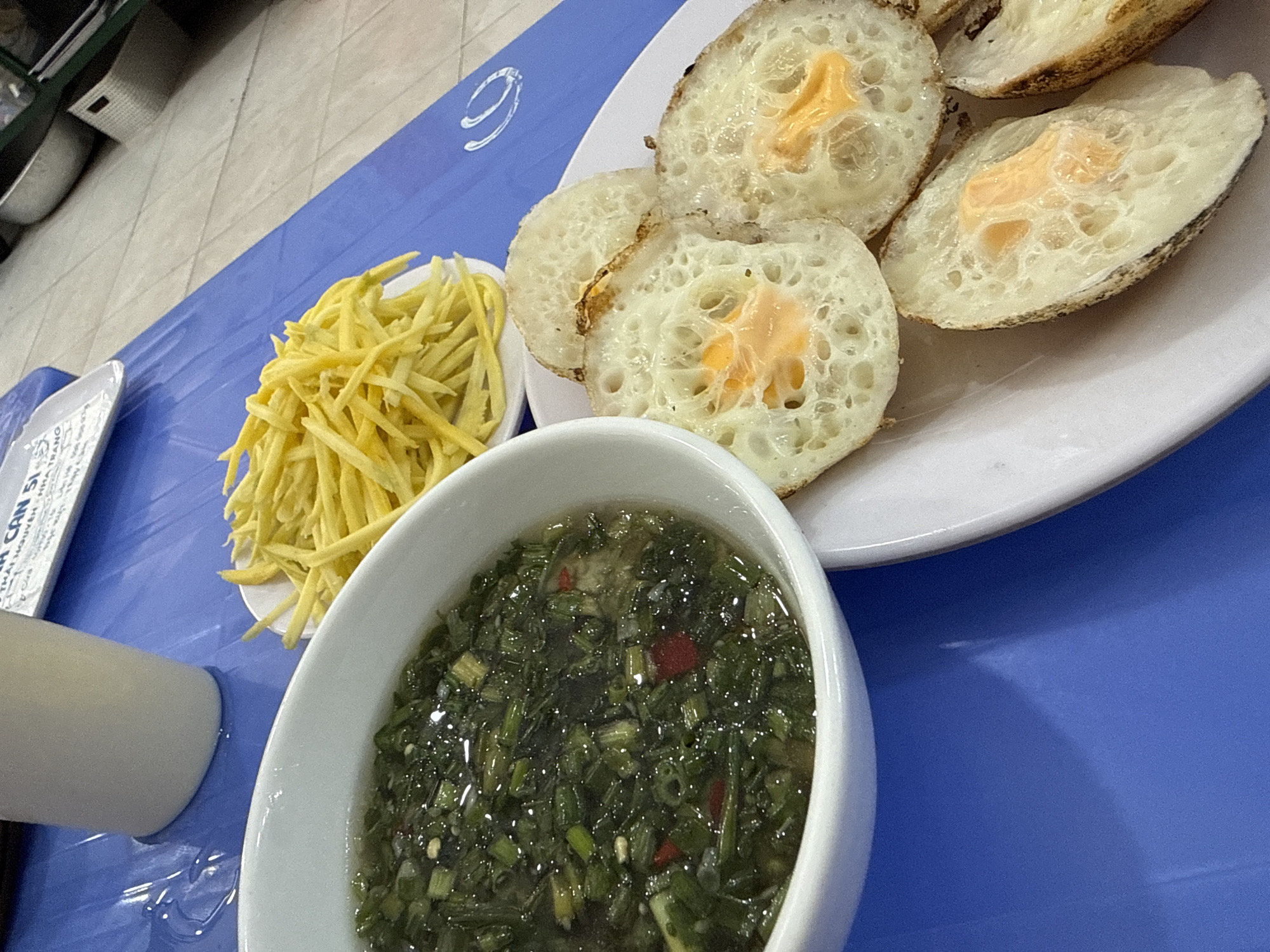 In Tay Nha Trang Ward, Vietnam, a close-up, high-angle shot captures a food spread on a blue plastic-covered table. The scene is illuminated by overhead lighting, creating reflections on the table's surface. In the foreground, a white bowl contains a dark green, chopped scallion-based sauce with visible red chili pieces. To the right, a white plate holds several small, round, golden-brown pancakes, each with a bright orange yolk filling. A small white dish filled with finely julienned yellow daikon radish sits behind the bowl of sauce. In the background, a white paper placemat with text is visible, as is a white ceramic cup. The overall setting suggests an outdoor or casual dining environment.