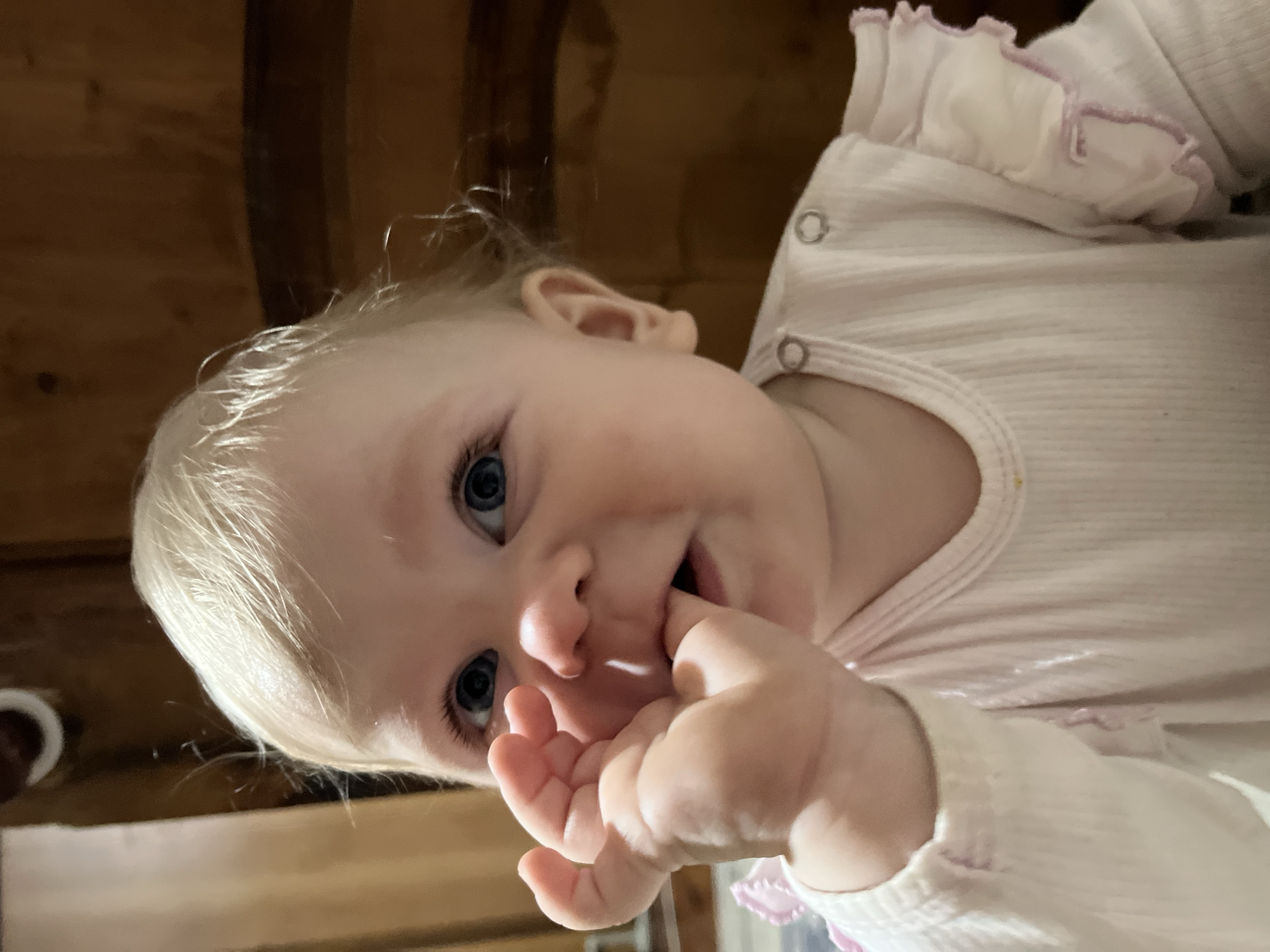 A baby with fair skin, blonde hair, and blue eyes is facing the camera with its fingers in its mouth. The baby is wearing a light-colored, long-sleeved garment with ruffle details around the neckline. The background appears to be a wood paneled surface. The location Vesyoloye, Russia is not related to the image.
