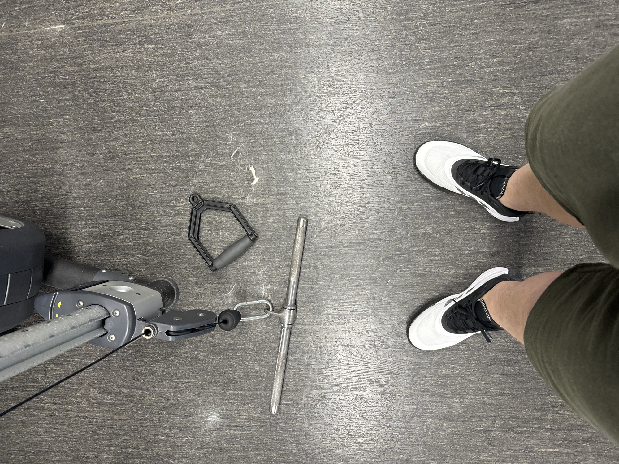 The image shows a view looking down at a section of what appears to be a gym. A cable machine is visible on the left side of the frame, with two different handle attachments, one triangular and one T-shaped, lying on the floor next to it. A person is standing in the frame on the right, wearing olive green shorts and white and black athletic shoes. The floor is a gray color with a textured surface. There is no information that can be used to discern if this image was taken in Varna, Bulgaria.
