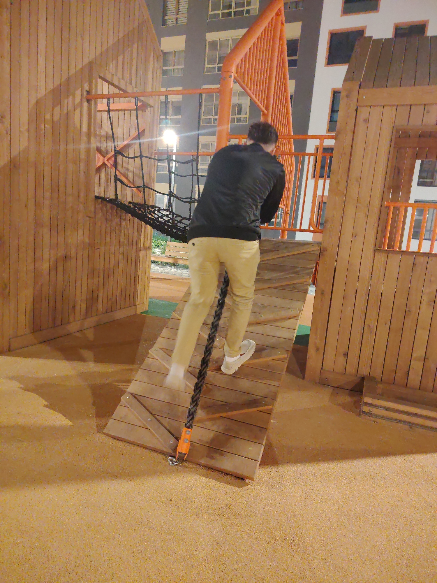 A person is climbing a rope ladder that is part of an outdoor playground structure. The playground is made of wood and has orange metal accents. The ground is covered in a soft, sandy material. In the background, there is a multi-story apartment building with many windows. The scene is lit by artificial lights, suggesting it is nighttime or dusk. The person is wearing a black jacket, khaki pants, and white sneakers. Their motion suggests they are actively ascending the ladder. This scene is located in Yekaterinburg, Russia.
