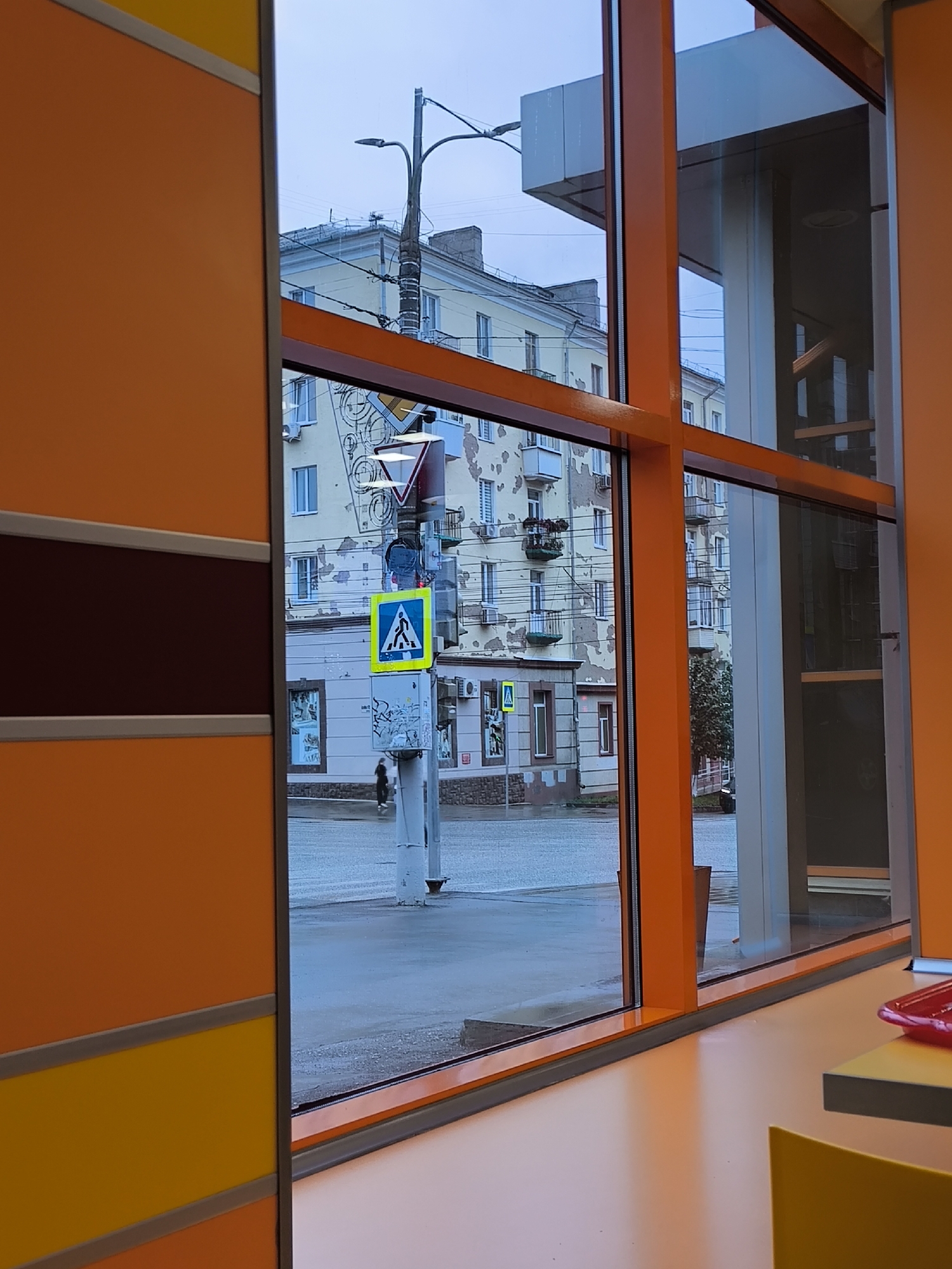 This image captures a view through a modern, orange-framed window, likely from an interior space looking out onto a city street. The exterior scene is dominated by overcast, cool-toned lighting, suggesting a cloudy or twilight day.
In the foreground, the vibrant orange of the window frame and interior walls creates a stark contrast with the muted colors of the outside. The window itself is large and multi-paneled, offering a panoramic view.
The street scene features several multi-story buildings with weathered facades, exhibiting peeling paint and balconies. A pedestrian crossing is clearly marked with a blue and white sign depicting a walking figure. A single pedestrian, a dark silhouette, is visible walking on the wet pavement near the crossing. Streetlights and utility poles are present, along with some greenery, possibly trees, in the distance. The ground appears wet, indicating recent rain or damp conditions.
The image is likely taken in Izhevsk, Russia, as the architectural style of the buildings and the presence of typical Russian street signs are consistent with urban environments in that region. The overall impression is one of a typical urban street scene observed from a contemporary interior space.