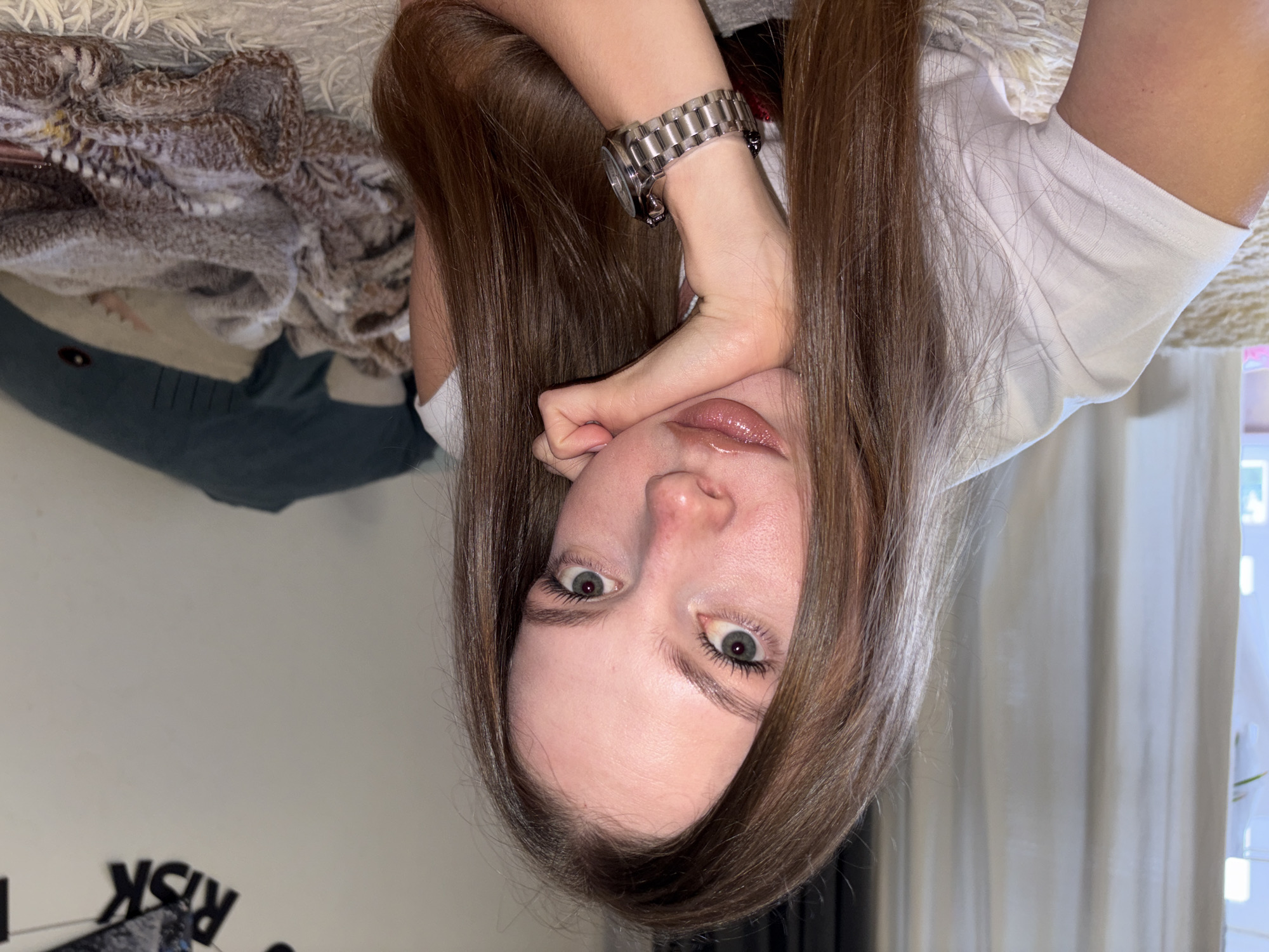 This image, taken in the city of Perm, Russia, presents a close-up portrait of a young woman. She is positioned on her side, with long, straight, brown hair framing her face and flowing around her shoulders. Her light blue-green eyes are wide open, looking directly into the camera, and her lips are full and coated with a glossy, light pink or nude product. Her left hand is raised, supporting her head with fingers gently curled beneath her chin. She wears a plain white short-sleeved t-shirt, and a silver-colored metal link watch is visible on her right wrist. The background includes a dark teal plush shark toy to her left, and a textured, crumpled blanket with a brown and cream pattern behind her head and right shoulder. In the upper left corner, partial black lettering spelling "RISK" is visible on a light-colored wall or surface. The scene is illuminated by soft, ambient light.