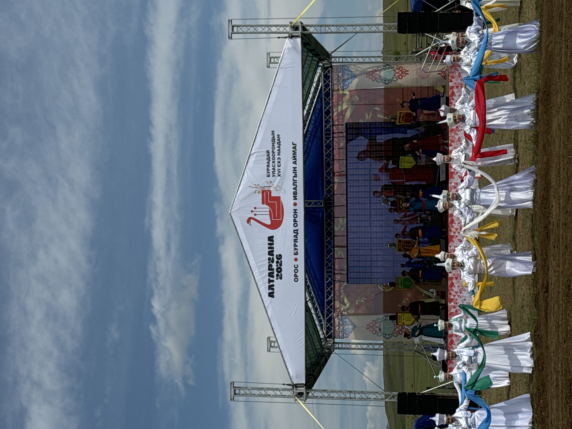 An outdoor stage setup is visible, featuring a white canopy with Cyrillic text and a red graphic of a bird. The text translates to "ALTARGAHANA 2026" and indicates the event is in Orongoi, Buryatia, Russia, specifically for the XVI Naadan. A group of individuals, appearing to be performers dressed in traditional white costumes and hats with colorful sashes, are positioned in the foreground, some holding and extending long, flowing sashes. Behind them, on the stage, other performers are visible, some standing and others engaged in what appears to be a traditional dance or performance, set against a backdrop displaying various images and patterns. The stage is supported by metal scaffolding and is set on a grassy, uneven terrain under a partly cloudy blue sky.