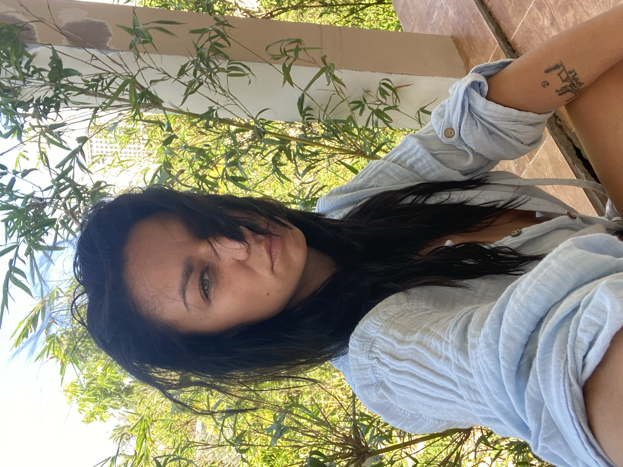A woman is pictured in a medium shot, seated outdoors with foliage visible in the foreground and background. The location is potentially Mũi Né, Vietnam based on the surroundings. She is wearing a light blue button-down shirt, and a tattoo is visible on her right arm. Her dark hair falls across her face, and she is looking directly at the camera. The environment appears to be a patio or balcony area with plants.