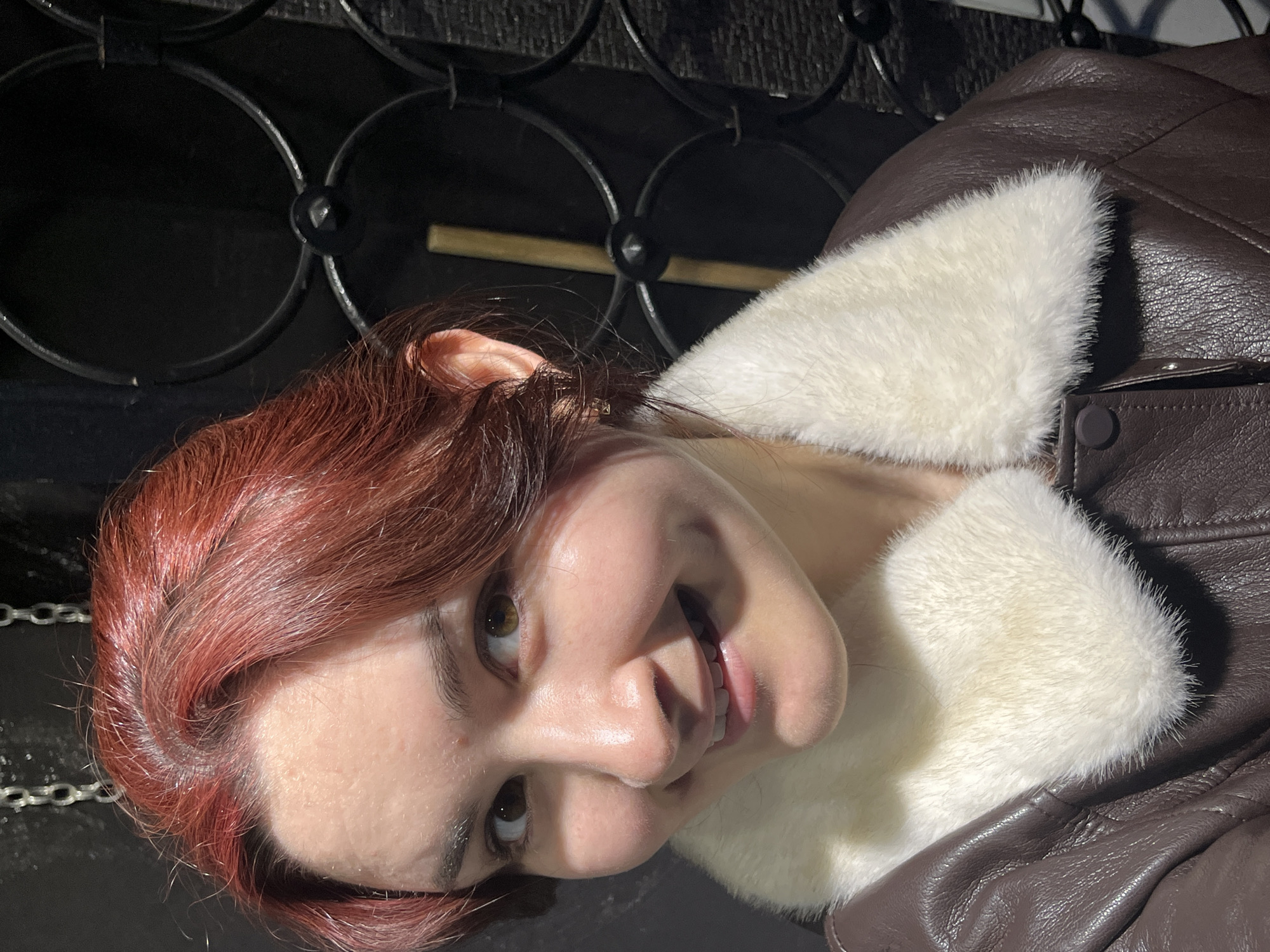 A young adult female is depicted in a frontal, waist-up portrait, looking directly at the camera with an open-mouthed smile, revealing teeth and dimples on both cheeks. She has red-dyed hair, styled away from her face, and brown eyes. Her skin tone is light, with some minor blemishes visible on her forehead. She is wearing a dark brown, possibly leather or faux-leather, jacket with a prominent cream-colored faux fur collar. A single dark brown button is visible on the jacket's front closure. A small, light-colored stud earring is visible in her right earlobe. The background is dark, featuring a metallic chain hanging vertically in the upper left. To the right, an ornate dark metal grate with interconnected circular and curved designs is present. Partially visible behind the grate, to the right of the subject's head, is a vertical light-colored wooden or metallic rod. The subject is well-lit from the front, contrasting with the dimly lit background. The specific location within Neftekamsk, Russia, cannot be determined from the visual information provided in this image.
