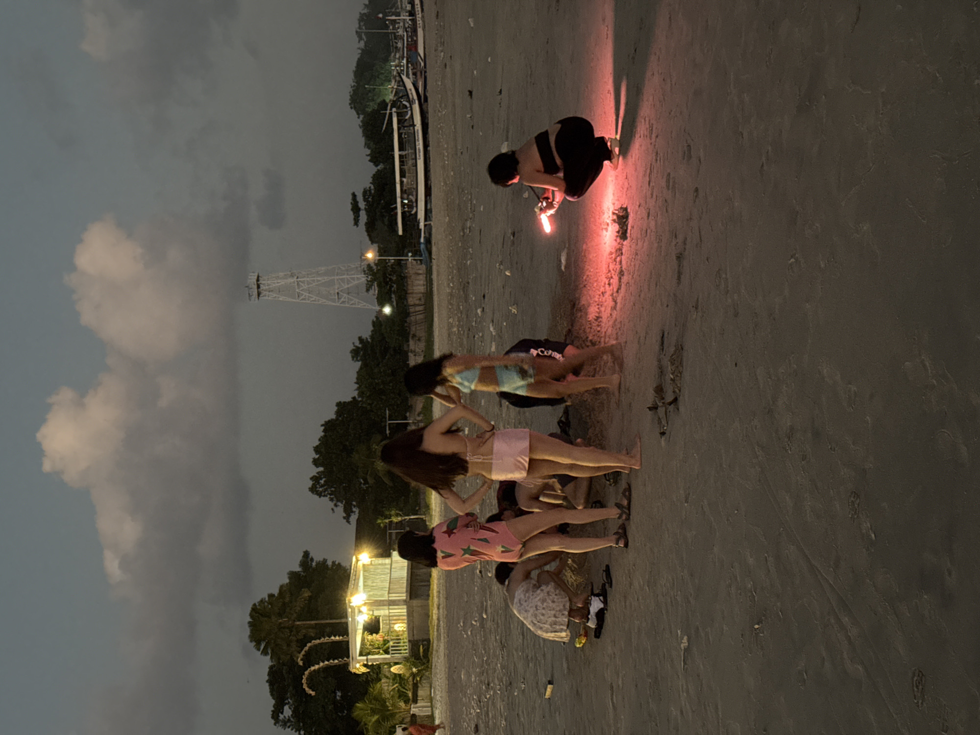 This is a medium-shot, eye-level, outdoor photograph taken at dusk on a sandy beach in Kuta, Indonesia. Several individuals, appearing to be young women, are gathered on the beach. In the foreground, a person is crouched down, holding a bright red flare that illuminates the sand around them. To the left of this individual, another person is standing, facing away from the camera. Further to the left, three other individuals are interacting in a group, with one appearing to be crouching.
In the mid-ground, several buildings with lit lamps are visible, suggesting a beachfront establishment or restaurant. Beyond the buildings, trees line the horizon. A tall, white, lattice-like tower is prominent in the background, a distinctive landmark. Several boats are moored on the water in the distance. The sky is overcast with dark clouds, indicating an impending or ongoing weather event. The overall scene depicts a social gathering on a beach at twilight.