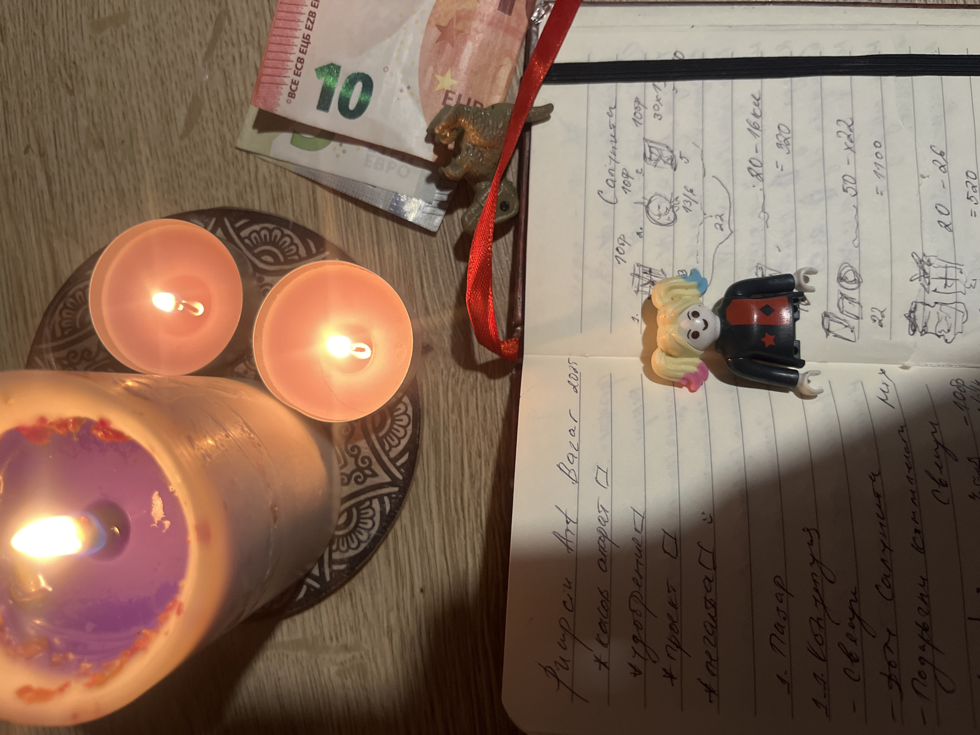 An overhead shot captures a cozy, illuminated scene centered around an open notebook and lit candles on a wooden surface. Three lit candles are visible: one large, thick candle with a purple center and white wax, and two smaller orange tealight candles, all resting on a decorative black and white patterned coaster or plate.
An open notebook, bound with a black elastic band and marked with a red ribbon, dominates the right side of the frame. The notebook contains handwritten text in Cyrillic script, some numbers, mathematical calculations, and small doodles. Visible words include "Art Bazar 2005," "касов апарат" (cash register), "проект" (project), "Пазар" (Market), "Свещи" (Candles), and "Санктум" (Sanctum). Calculations like "20-16 = 320" and "50-x22 = 1100" are present. A small figurine resembling Harley Quinn, with blonde pigtails and a red and black outfit, rests on the notebook's page. Another small, brown animal figurine is perched near the spine of the notebook, partially obscured by the red ribbon bookmark.
To the upper left, several euro banknotes are partially visible, including a prominent 10 euro note displaying "10," "ЕВРО," and "BCE ECB ЕЦБ EZB EKP." The ambient lighting suggests an evening or nighttime setting, with the candle flames casting warm glows. The overall arrangement creates an atmosphere of introspection, planning, or a quiet ritual.