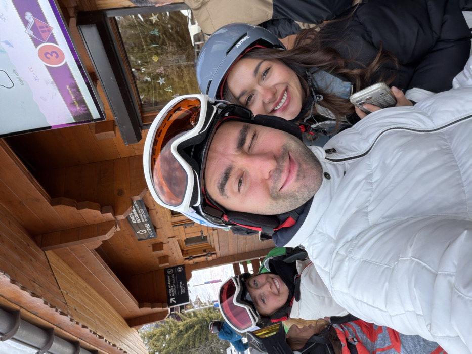 A close-up selfie captured at a ski resort in Les Allues, France, features three individuals in winter ski gear with additional people partially visible in the background. The primary subject in the foreground, a male, occupies the center-left, wearing a white quilted ski jacket, a black ski helmet with red earpads, and white-framed ski goggles with orange lenses pushed up onto the helmet. He smiles with his mouth closed and winks with his left eye. To his right, a female wearing a dark ski jacket and a grey "Atomic" brand ski helmet with a red interior lining smiles toward the camera. She holds a light-colored mobile phone with a textured case in her right hand, positioned near the male's midsection. To the male's left, partially obscured, is another female wearing a white ski jacket and a black ski helmet with red-framed ski goggles pushed up; she is also smiling.
The background features a multi-story wooden building constructed with horizontal planks and decorative carved wood elements. A dark grey gutter system is visible along the roofline. Mounted on the building's facade are several signs: one sign to the left indicates "ACCÈS TÉLÉCAGE DU GOLF ET ACCÈS CHALILIFT ACCESS" with an arrow, and "TOILETTES" with a toilet icon. Another sign to the right states "ACHAT & DE COMMA" / "RAPID PASS." Above a dark roller shutter, a flat-screen monitor displays weather information in French, including "Soleil voilé," temperatures "3°C - 4°C," and "Risque d'avalanche" at level "3." A window or door panel is adorned with festive star and tree cutouts. A snowy ski slope and evergreen trees are visible further in the background, with a person in a blue jacket and ski helmet partially seen on the slope to the far left.