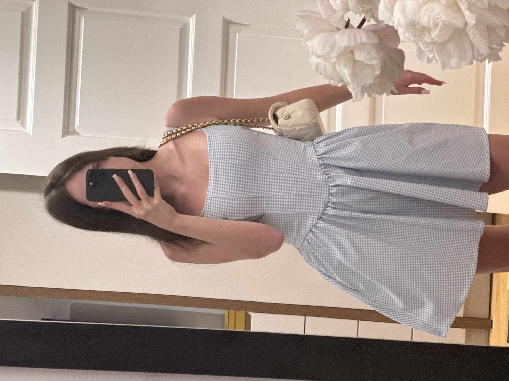 A young woman is depicted taking a mirror selfie in an indoor setting. She holds a black smartphone to her face, obscuring her eyes and part of her nose, with her light-colored, manicured nails visible around the device. Her long, dark hair falls around her shoulders.
She is wearing a strapless dress featuring a light blue and white gingham pattern, with a gathered or ruffled bodice and a full skirt. A small, quilted, off-white or cream-colored handbag with a gold chain strap is slung over her right shoulder.
The background reflected in the mirror shows white paneled doors, suggesting a clean, classic interior design. In the upper right portion of the image, several large, fluffy white flowers, resembling peonies, are visible, adding a decorative and soft touch to the scene. Below the mirror, a dark, horizontal object is partially visible, possibly part of a console or shelf. The lighting is bright and even throughout the image, indicating a well-lit indoor environment. There is no visible text.