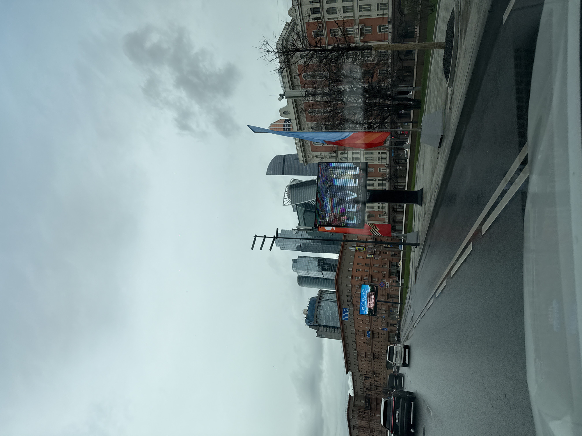 The image depicts a street scene in Moscow, Russia, on an overcast day. The view appears to be taken from inside a vehicle moving along a road. The road is wet, suggesting recent or ongoing rain. Along the roadside, there are buildings of varying architectural styles, including older brick structures and modern skyscrapers.
Russian flags are visible along the street. A digital billboard displays the word "LEVEL," along with abstract graphics. Several cars are present on the road.