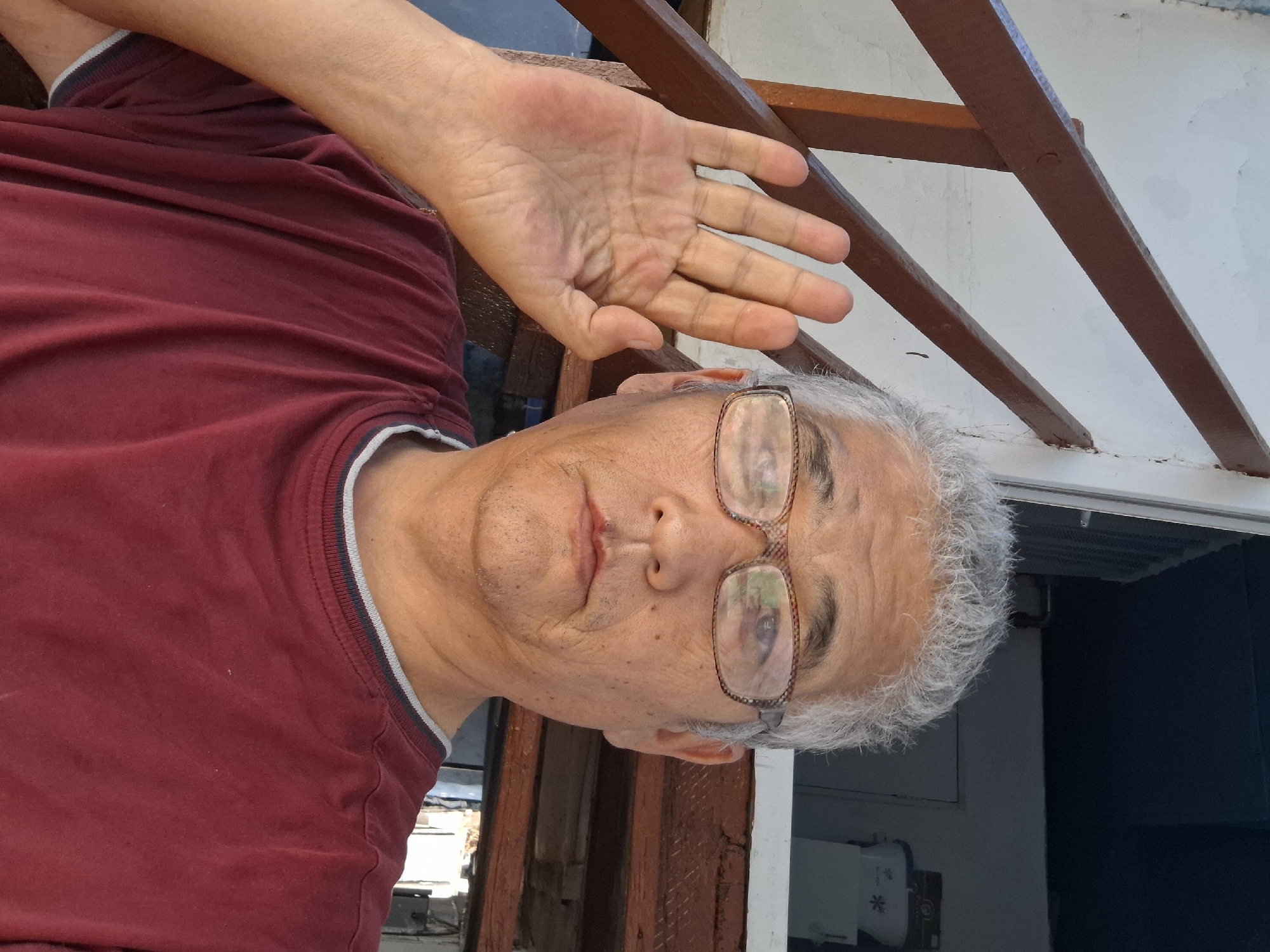 A middle-aged man with graying hair and glasses is taking a selfie. He is wearing a maroon t-shirt and has his left hand raised to his ear. The background appears to be outdoors, possibly on a porch or balcony, with wooden railings and a white wall. The environment suggests a residential area. The image does not provide specific geographical markers to pinpoint a location within Almaty, Kazakhstan.