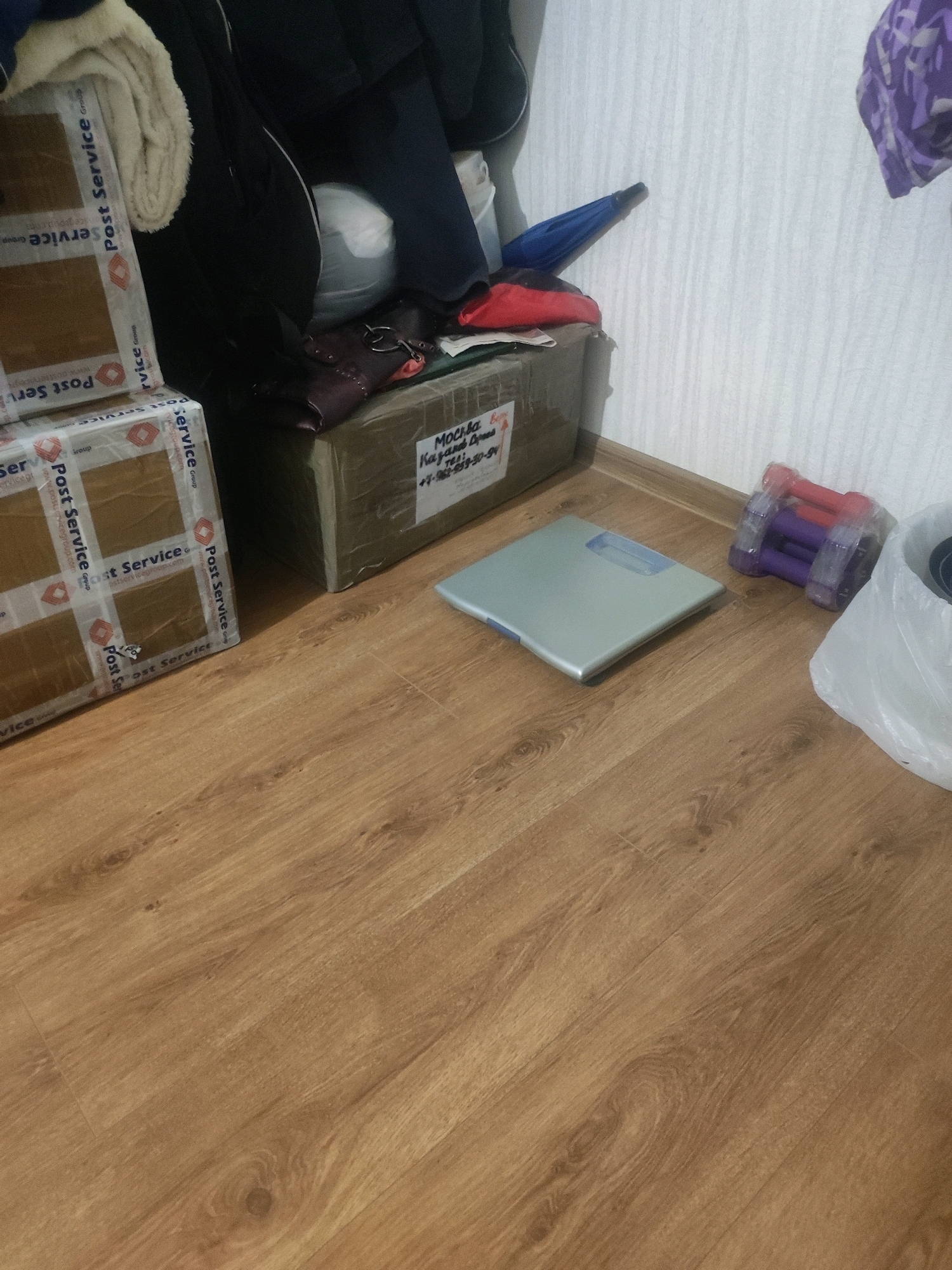 The image shows a cluttered floor in what appears to be a storage area or a room in Moscow, Russia. The floor is made of light brown laminate flooring with a wood grain pattern. On the floor, there are several objects, including two cardboard boxes sealed with brown tape and marked with "Post Service Group" logos, indicating they may have been shipped. One of the boxes has a handwritten label in Russian that reads "Москва" (Moscow) and other text that appears to be an address or recipient information.
Next to the boxes is a silver digital bathroom scale with a blue display. To the right of the scale, there is a package containing several small dumbbells, some of which are purple and red. Further to the right, a large white plastic bag is partially visible, and behind it, a purple and white patterned fabric item is draped over something out of frame. In the background, against a white textured wall, a blue folded umbrella is leaning, and dark clothing or bags are stacked. The overall impression is one of a disorganized space with items that have likely been stored or are awaiting transport.