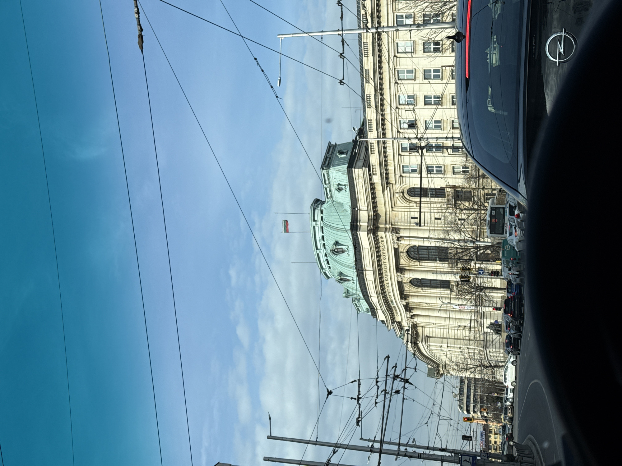 The image depicts a view from inside a vehicle, likely in Sofia, Bulgaria. The Alexander Nevsky Cathedral is visible, recognized by its distinctive green dome and the Bulgarian flag atop it. Streetcar wires crisscross the sky in the foreground. The vehicle in which the photo was taken appears to be an Opel, as indicated by the logo on the rear of the vehicle. The photo was taken during daylight with a mix of blue sky and clouds, with other vehicles ahead on the road, suggesting traffic.
