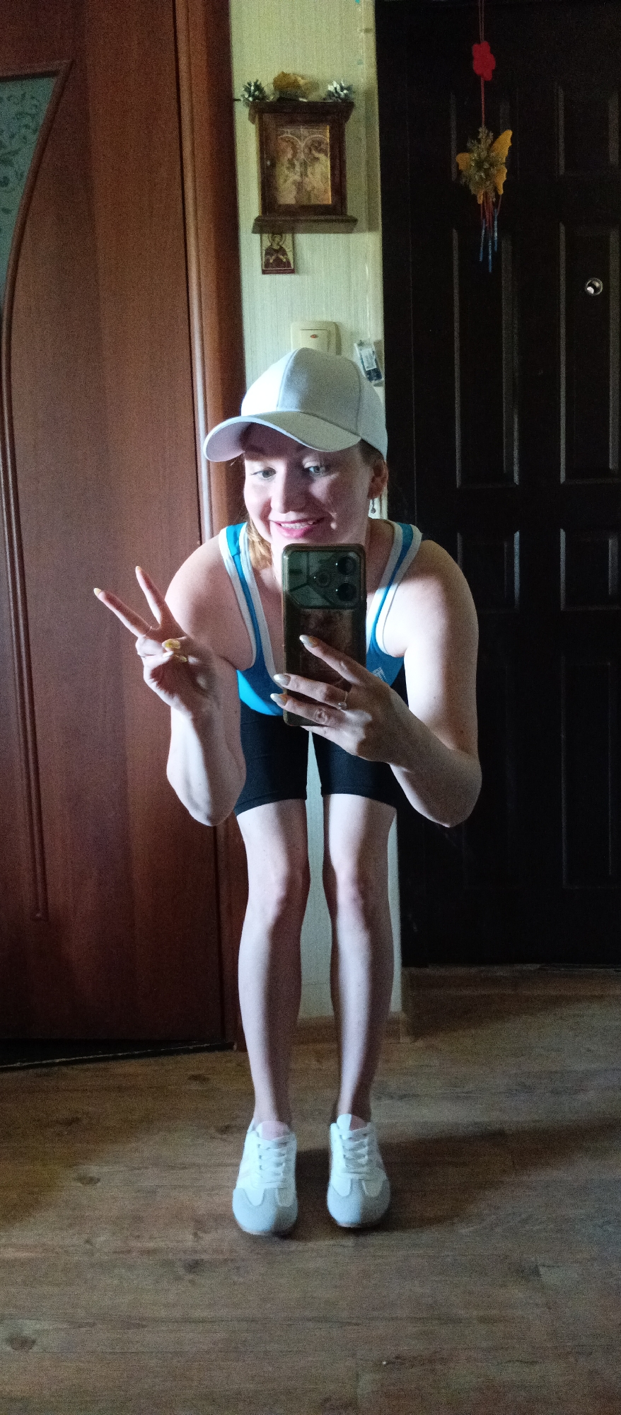 A full-body, medium-shot photograph shows a woman inside a home. She is in an Unknown city and state. She is bending forward slightly and facing the camera while holding a smartphone in her right hand, angled toward her face, as if taking a selfie. Her left hand is raised, making a "peace" sign. The woman has fair skin and reddish blonde hair. She wears a white baseball cap, a light blue athletic tank top with white trim, black athletic shorts, and white sneakers. The woman appears to be in her late 30s or early 40s.
The background includes a wooden door on the left and a dark brown door on the right. A small wooden icon cabinet hangs on the wall between the doors, and a hanging decoration with a butterfly and red flower design is visible to the right of the cabinet. The floor is made of wood planks. The light source is above the camera, creating shadows in the lower portion of the image.
