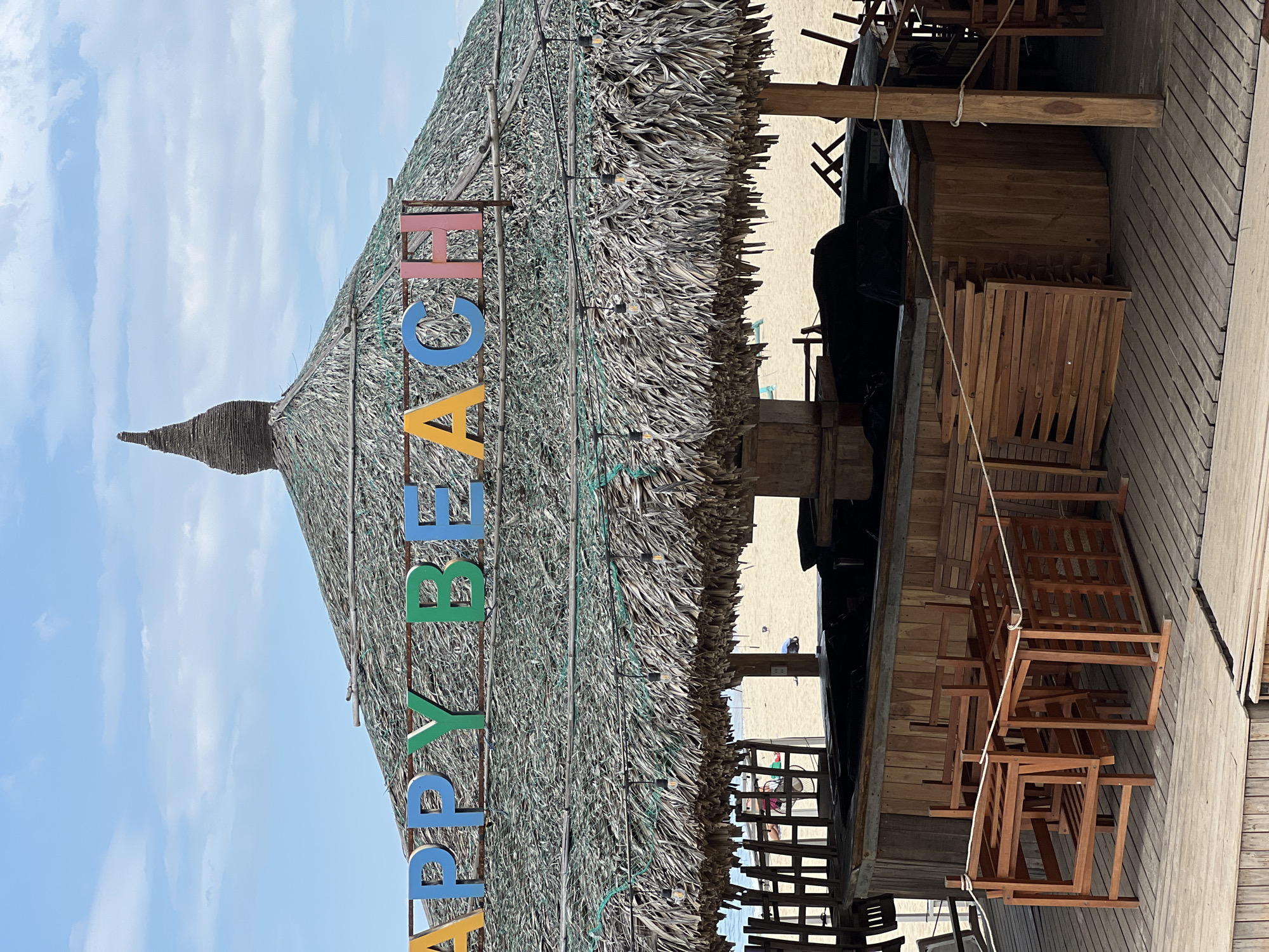 The image shows a beachside bar with a thatched roof in Nha Trang, Vietnam. The words "HAPPY BEACH" are displayed in colorful, block letters on the roof. Underneath the roof, a wooden bar counter is visible, with several wooden chairs stacked and tied with rope. The beach and the ocean can be seen in the background, with a few people scattered on the sand. The sky is partly cloudy. The overall scene suggests a casual, outdoor establishment on a sunny day.