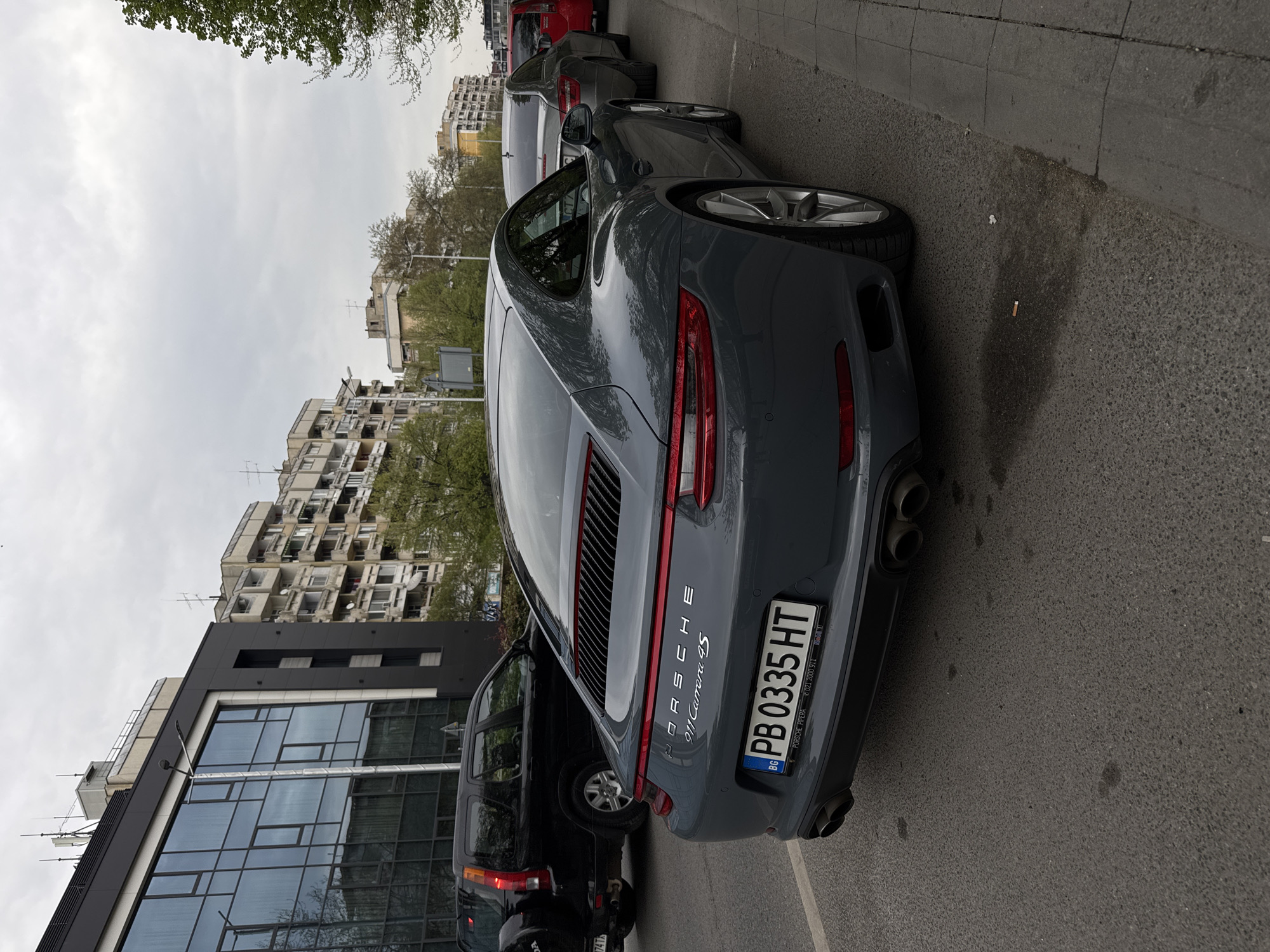 The image depicts the rear of a grey Porsche 911 Carrera 4S parked on a street. The license plate reads "PB 0335 HT" and indicates the car is registered in Bulgaria. To the left of the Porsche, a black SUV is parked. Behind the Porsche, other parked cars are visible. The background features a modern building with mirrored glass windows and several older residential buildings. The scene appears to be set on a typical street in an urban environment, most likely in the city of Plovdiv, Bulgaria.