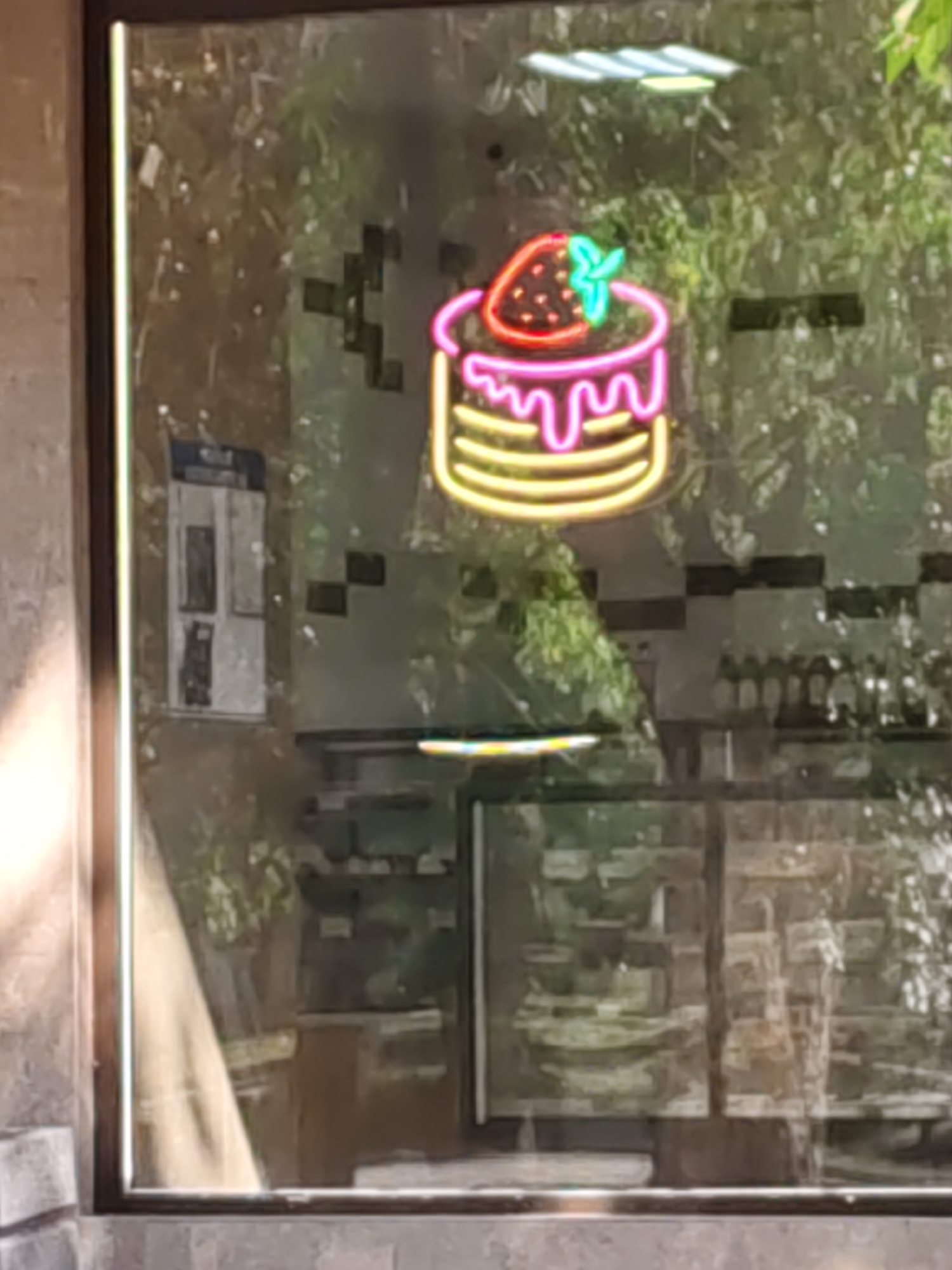 A neon sign shaped like a strawberry-topped cake is displayed on a window, possibly of a cafe or bakery. The cake has yellow layers and pink frosting dripping down the sides. The strawberry on top is red with black seeds and a green stem. The window reflects an outdoor environment with trees and sunlight. Inside the cafe, shelves stocked with bottles and what appear to be food items are visible. A flyer or poster is attached to the window.