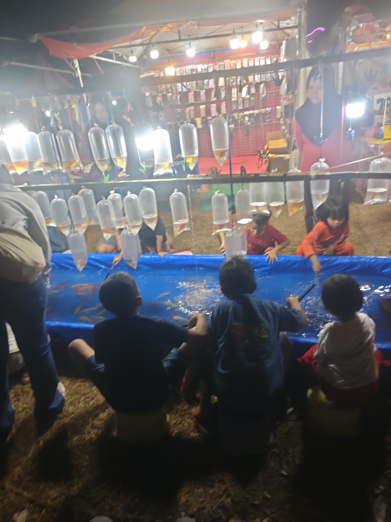In Subang, Indonesia, an outdoor night market features a fish-catching game. Children sit on the ground, attempting to scoop fish from a large blue pool using small nets. The pool is surrounded by a bright blue tarp. Above the pool, fish in clear plastic bags are suspended, presumably for sale or as prizes. The background shows stalls with merchandise, possibly phone accessories, and bright lighting illuminates the scene. A person in a hijab is visible behind the fish bags.