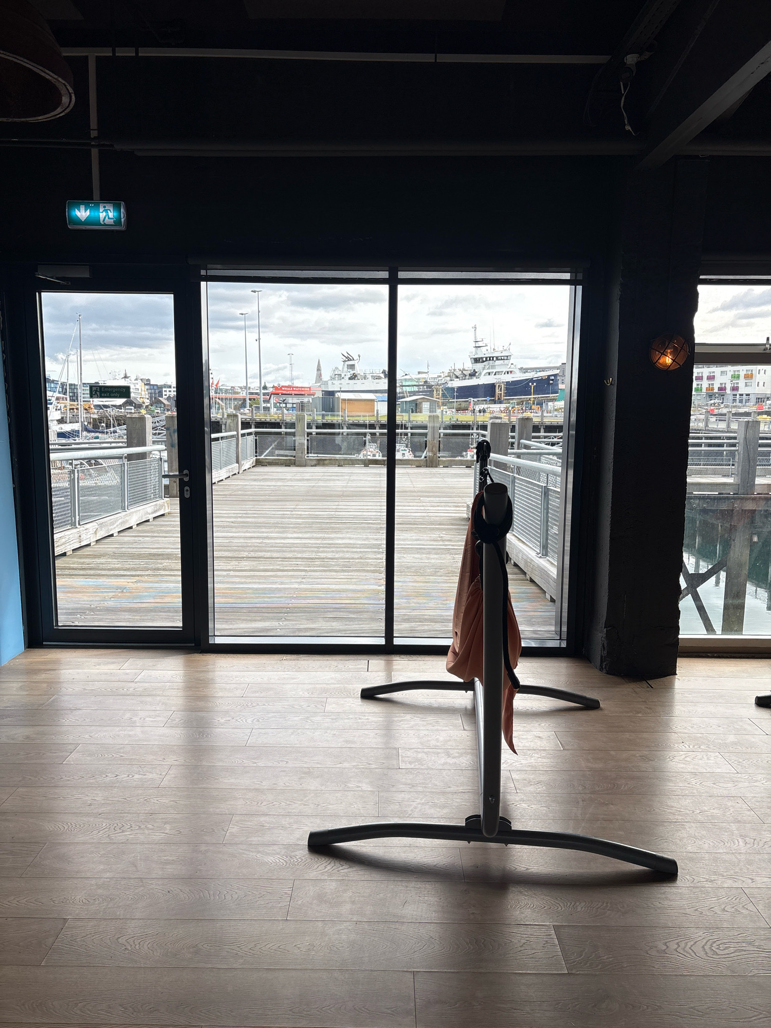 This is a full-frame, eye-level, outdoor shot taken within a building, looking out through large glass doors onto a wooden deck and a harbor in Reykjavik, Iceland.
The interior foreground is dominated by a light-brown wooden floor with visible plank lines and light reflections. In the center of the frame, a dark grey metal structure with curved legs stands on the floor. A light-orange garment, possibly a jacket or scarf, is draped over the vertical support of this structure. To the left, a green emergency exit sign with down arrows and a running person icon is mounted on a dark ceiling or wall.
The glass doors reveal a wooden deck area, made of weathered planks, that slopes slightly downwards towards the water. Metal railings with a mesh infill line the sides of the deck. Beyond the deck, a harbor is visible with several boats and ships docked, including a large white and blue vessel with "WHALE WATCHING" written on its side. The sky is overcast.
The overall atmosphere suggests a commercial establishment, likely a restaurant or cafe, located by the harbor.