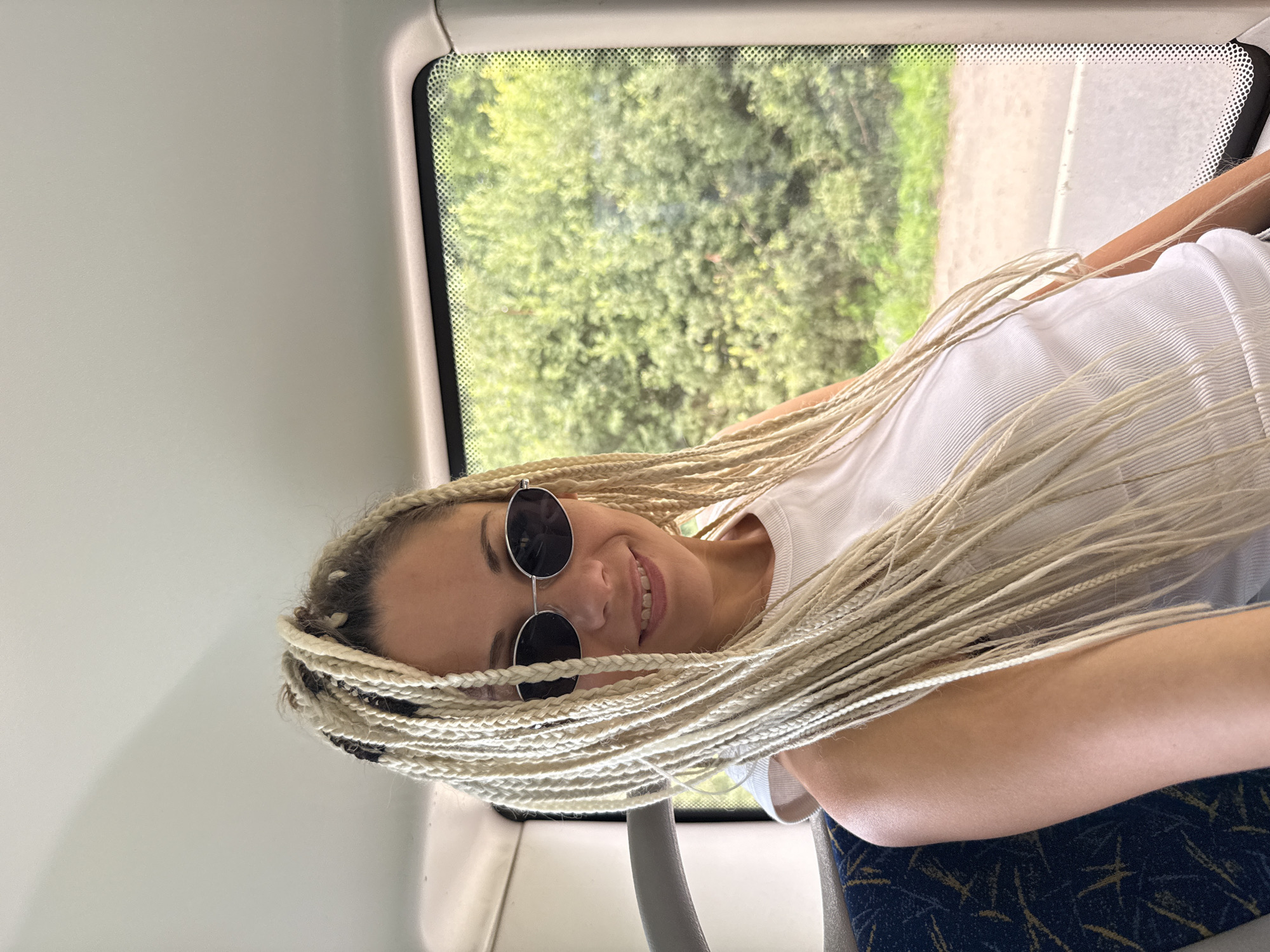 A young woman with blonde braids and sunglasses smiles while sitting in a vehicle, likely a bus or train, with a green, leafy exterior visible through the window. The interior of the vehicle shows a grey seat with a blue and yellow pattern and a white ceiling. The woman is wearing a white tank top.