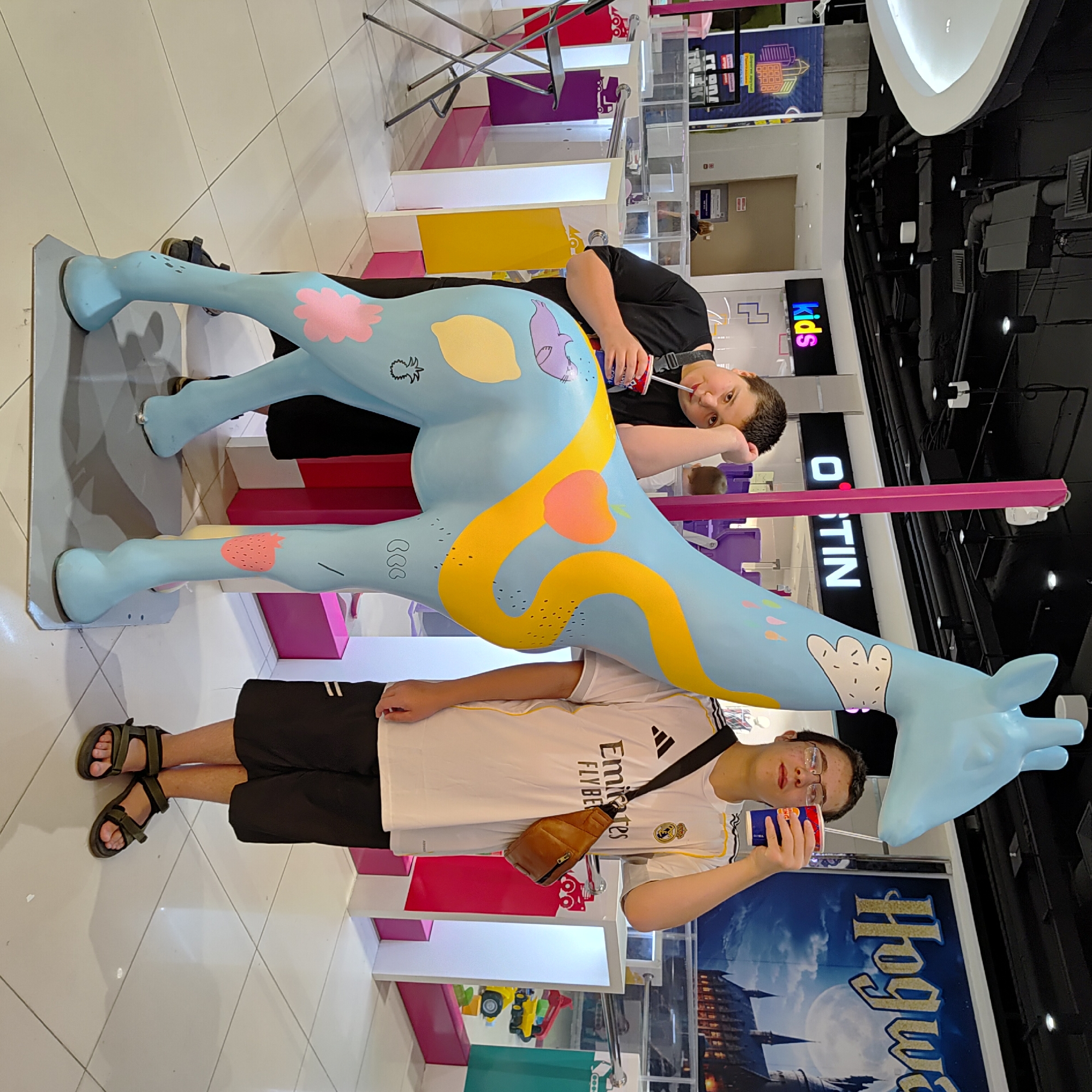Two young people stand in front of a large, blue, decorated giraffe statue in what appears to be a shopping mall. The person on the left, wearing a black t-shirt and black shorts, leans on the statue and drinks from a red cup with a straw. The person on the right, wearing a white t-shirt with black stripes and black shorts, holds a red and blue cup and looks towards the camera. Both individuals appear to be teenagers. The giraffe statue has yellow, pink, and white decorations, including fruit and animal shapes. In the background, there are store signs, including one that reads "kids" and another that says "Hogwarts." The floor is tiled with large white squares. There is no visible indication in the image that this location is in Ufa, Russia.