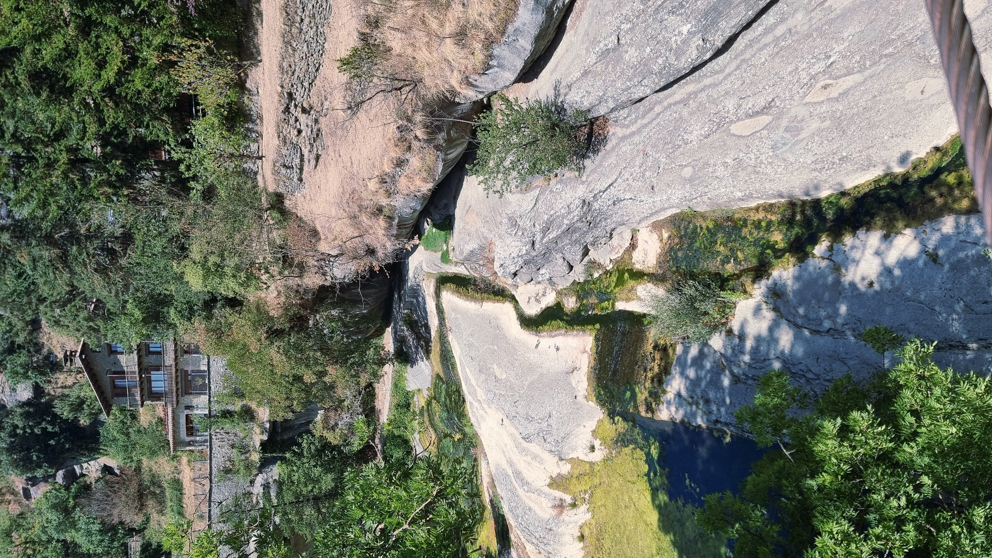 The image shows a scenic view of a natural landscape near Rupit i Pruit, Spain. A multi-story stone building with wooden balconies is situated on a hillside, surrounded by lush green trees and vegetation. Below the building, a shallow river flows over smooth, light-colored rocks, with patches of green algae or moss visible in the water. The terrain is rugged, with rocky outcrops and dry grasses on the slopes. The scene is bathed in bright sunlight, casting shadows on the ground and rocks.