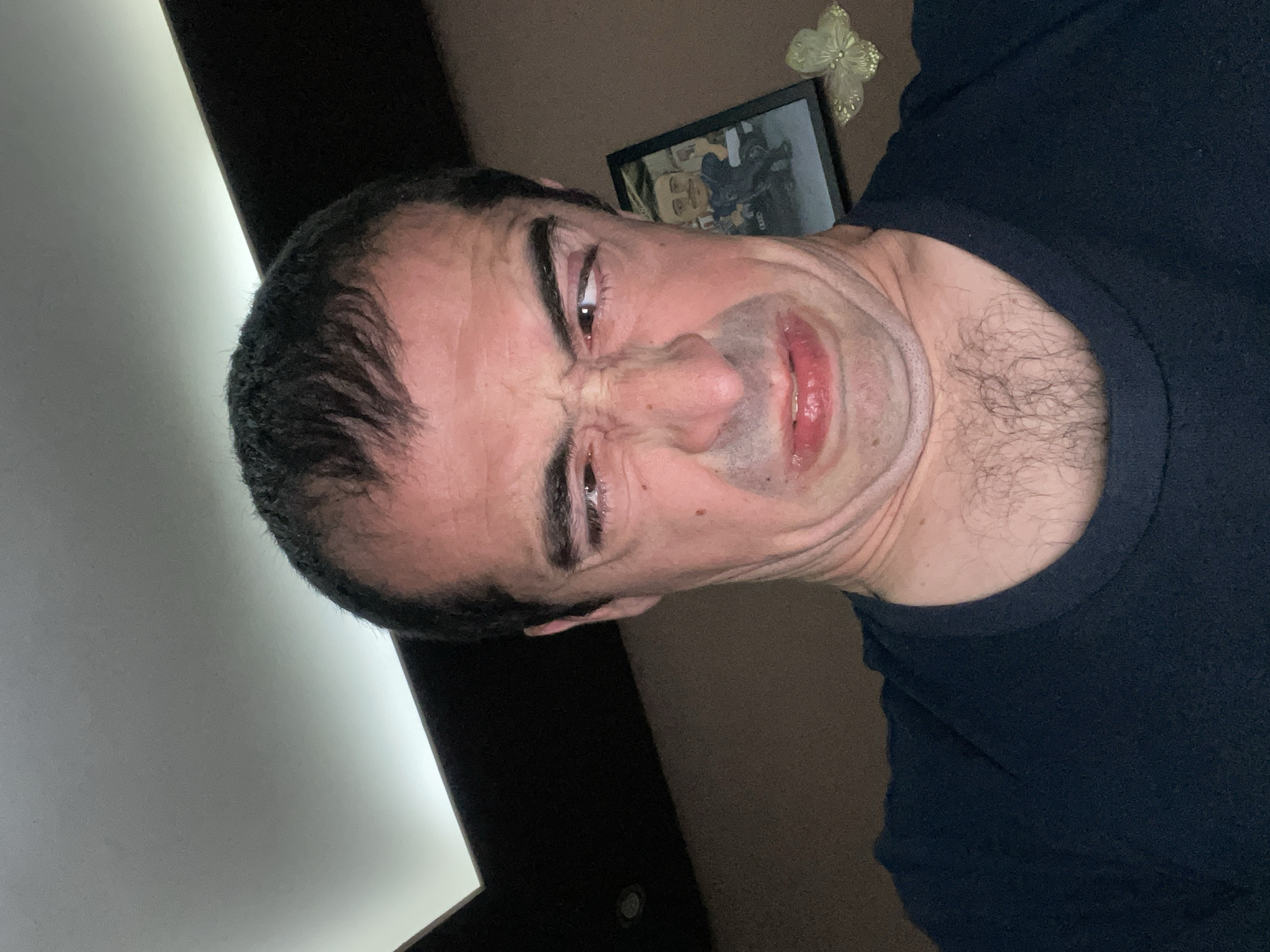 A selfie of a middle-aged man with dark hair and a grimaced expression. He is wearing a dark blue t-shirt. The background includes a white wall, a brown wall, a framed photograph, and a decorative butterfly. The location in Sofia, Bulgaria cannot be determined from the image.