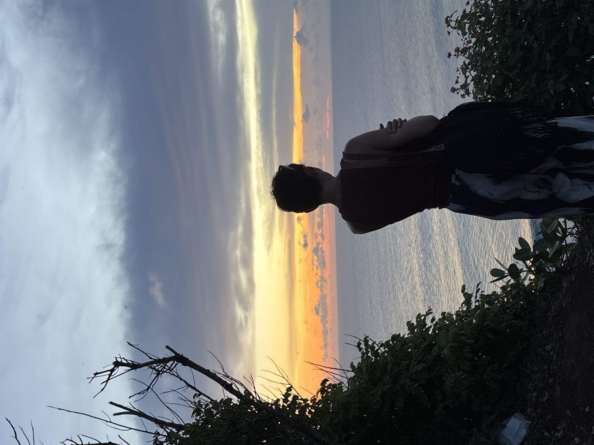 The image captures a sunset scene in Uluwatu, Indonesia. A person stands in the foreground, their back turned to the viewer, arms crossed. They wear a red top and patterned shawl, with visible green vegetation around them. The sky displays a mix of light blue, grey, white, orange, and yellow hues from the setting sun and clouds. The ocean shimmers in a similar color palette due to the sunset. Branches and plant life are silhouetted in the bottom of the frame, suggesting a cliffside location.